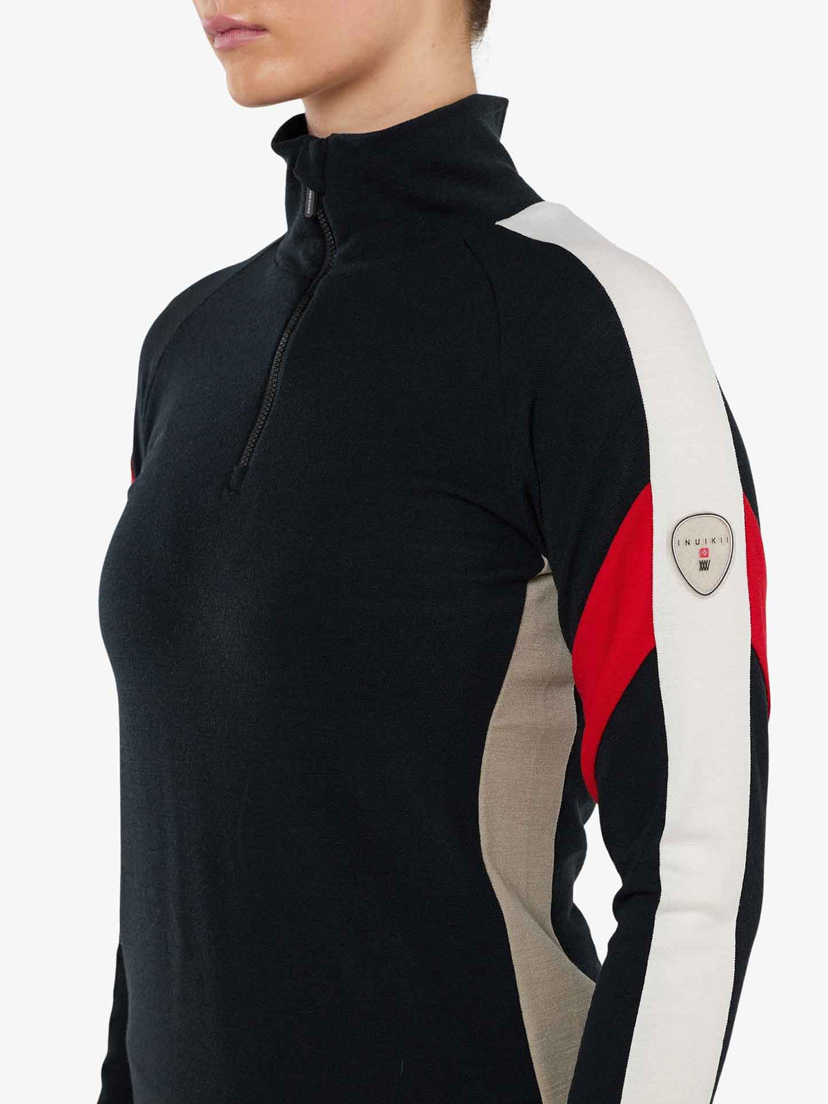 A person wearing the We Norwegians INUIKII + WN Zip Up Women, a black long-sleeve base-layer top with a high collar and a partial zipper. The sleeves feature red, white, and beige color blocks, accented by a small circular logo patch on the left arm. Part of an exclusive capsule collection, this stylish piece is perfect for any wardrobe. The background is plain white. The person's face is not visible.