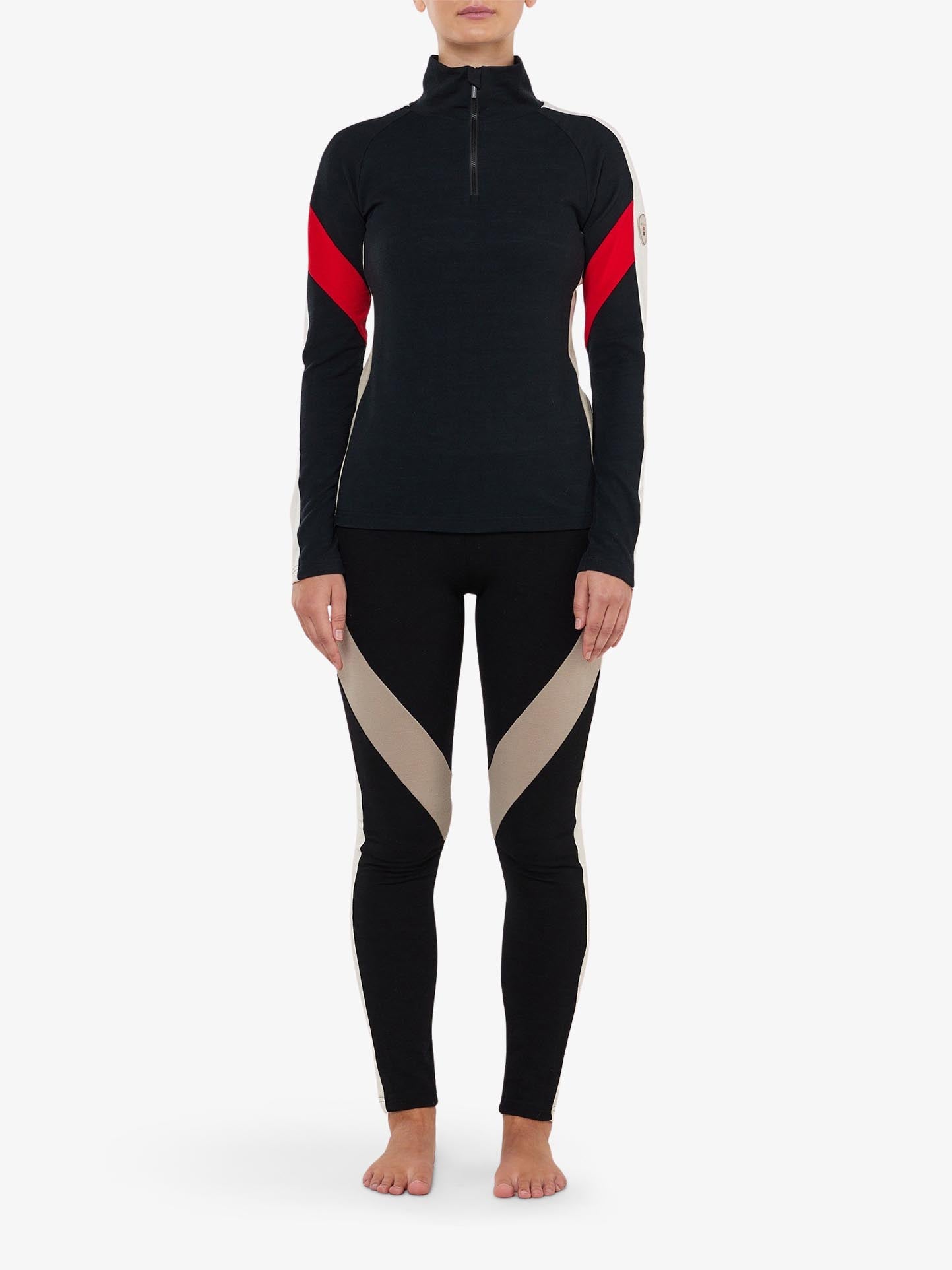 A person stands barefoot, wearing the sleek black "Inuikii+WN Leggings Women" by We Norwegians. The outfit is adorned with beige and red accents, making it perfect for ski fashionistas. The long-sleeved top has a front zipper, while the leggings feature diagonal beige stripes that resemble stylish legwear. The backdrop is a plain white canvas.