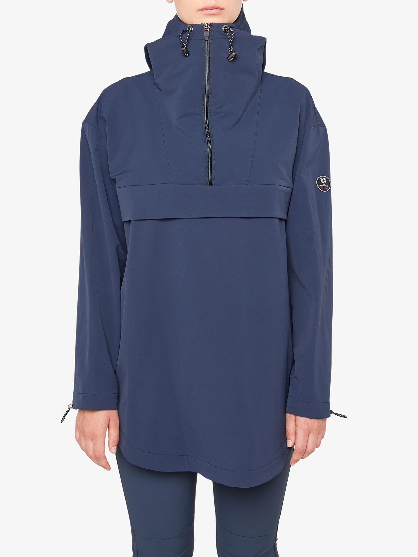 A person is wearing a loose-fitting, long-sleeved We Norwegians Beito Anorak Women in navy blue, boasting 4-way stretch and a high collar with a front zipper. The jacket features a horizontal seam across the chest and displays a small circular logo on the left sleeve. The person's hands are by their sides.