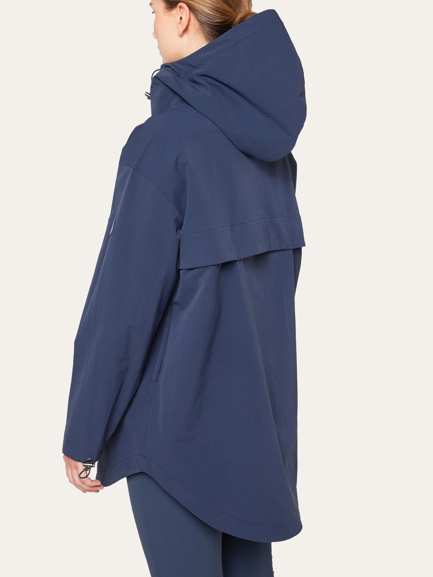 A woman with light brown hair wears the We Norwegians Beito Anorak Women in navy. The back view highlights its loose fit and vented yoke, reflecting classic Norwegian style. The background is plain and light-colored.