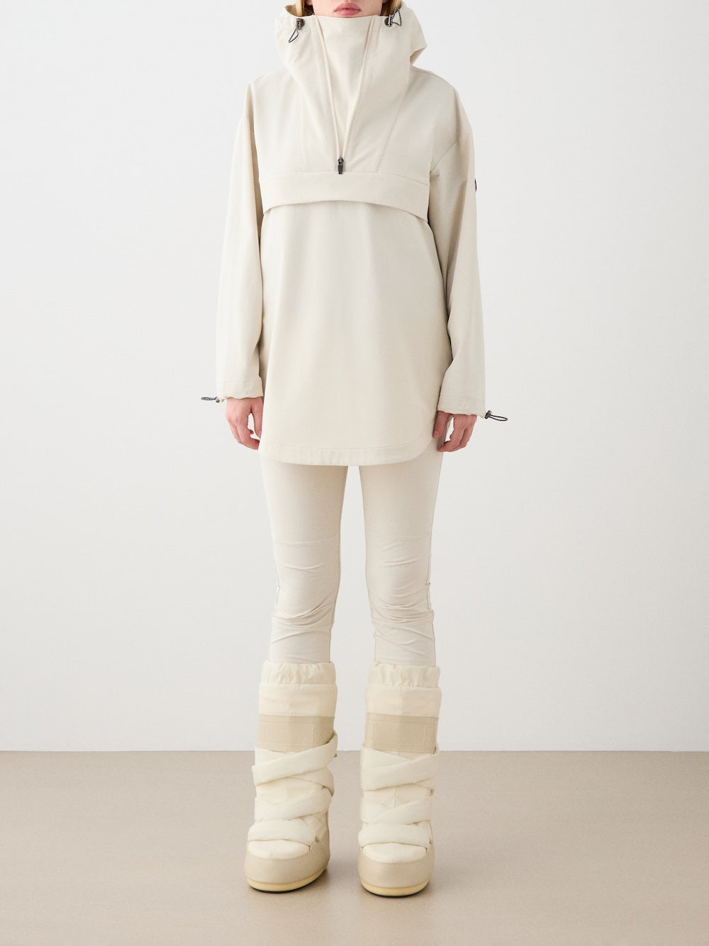 A person stands against a plain background in the We Norwegians Beito Anorak Women, with matching fitted pants and cream winter boots, channeling Norwegian outdoor style. The high collar of the jacket partially obscures their face.