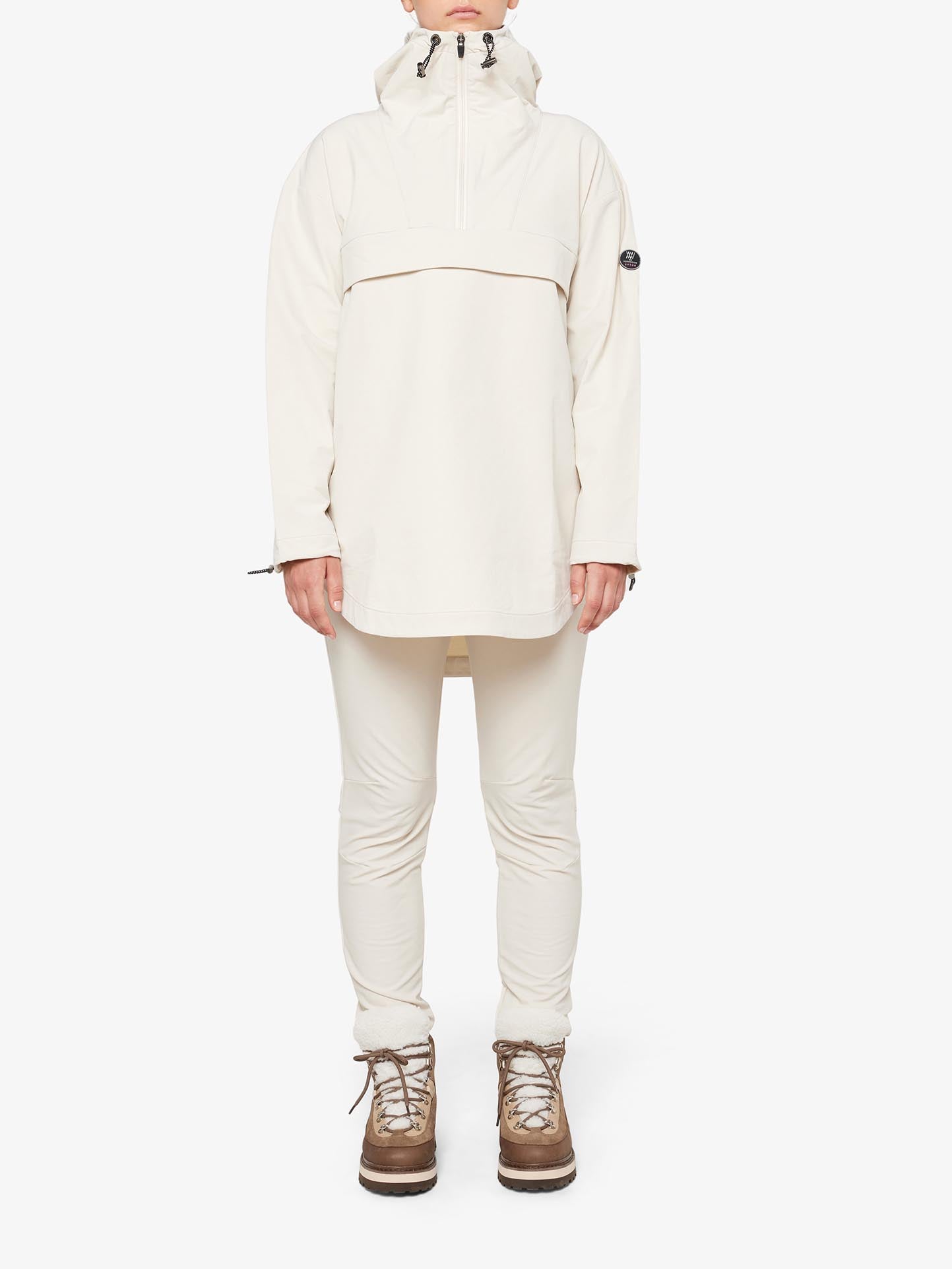 A person is wearing the Beito Anorak Women jacket by We Norwegians, which features long sleeves, a hood, and wool lining in beige. They are also dressed in matching beige pants and light brown hiking boots. The jacket includes a high collar with a zipper. The person is standing against a plain white background with their face not visible.