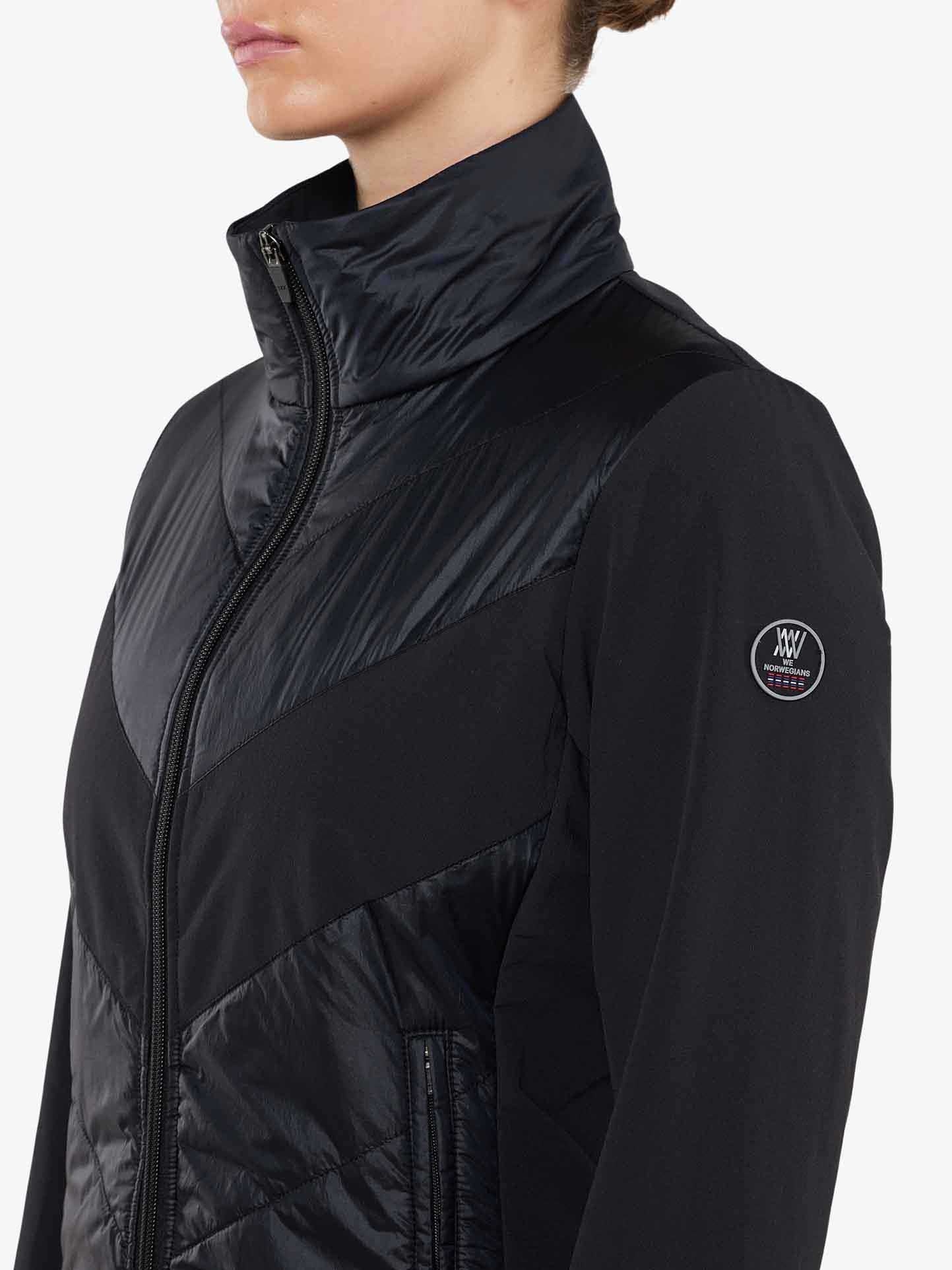 A person wears the We Norwegians Beito Jacket Women, featuring a high collar and a zippered front. The jacket boasts a shiny, quilted texture on the upper chest and shoulders, with a circular emblem on the sleeve that alludes to its cross-country skiing origins. The background is plain white.