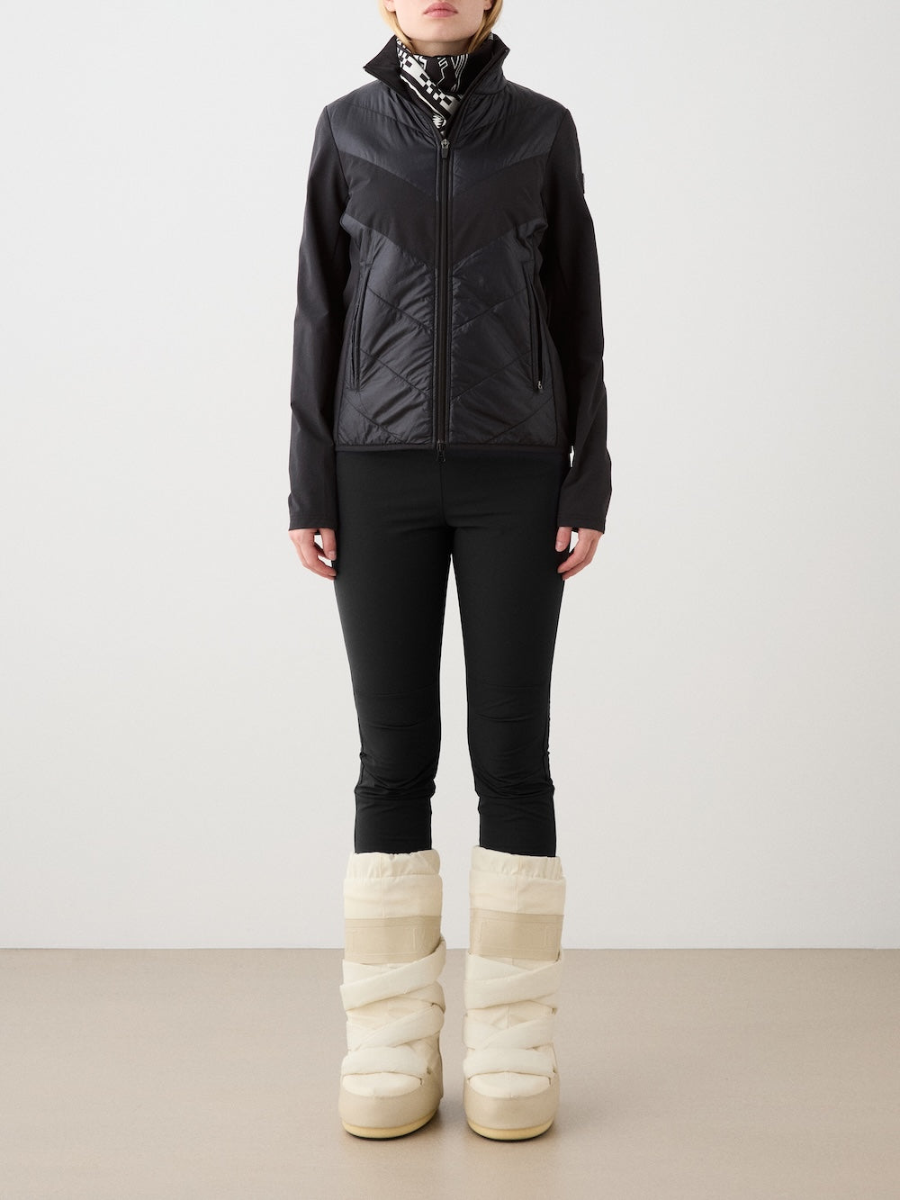 A person stands against a plain background wearing a black We Norwegians Beito Jacket Women, black leggings, a patterned scarf, and cream-colored padded winter boots, with their hands relaxed at their sides.