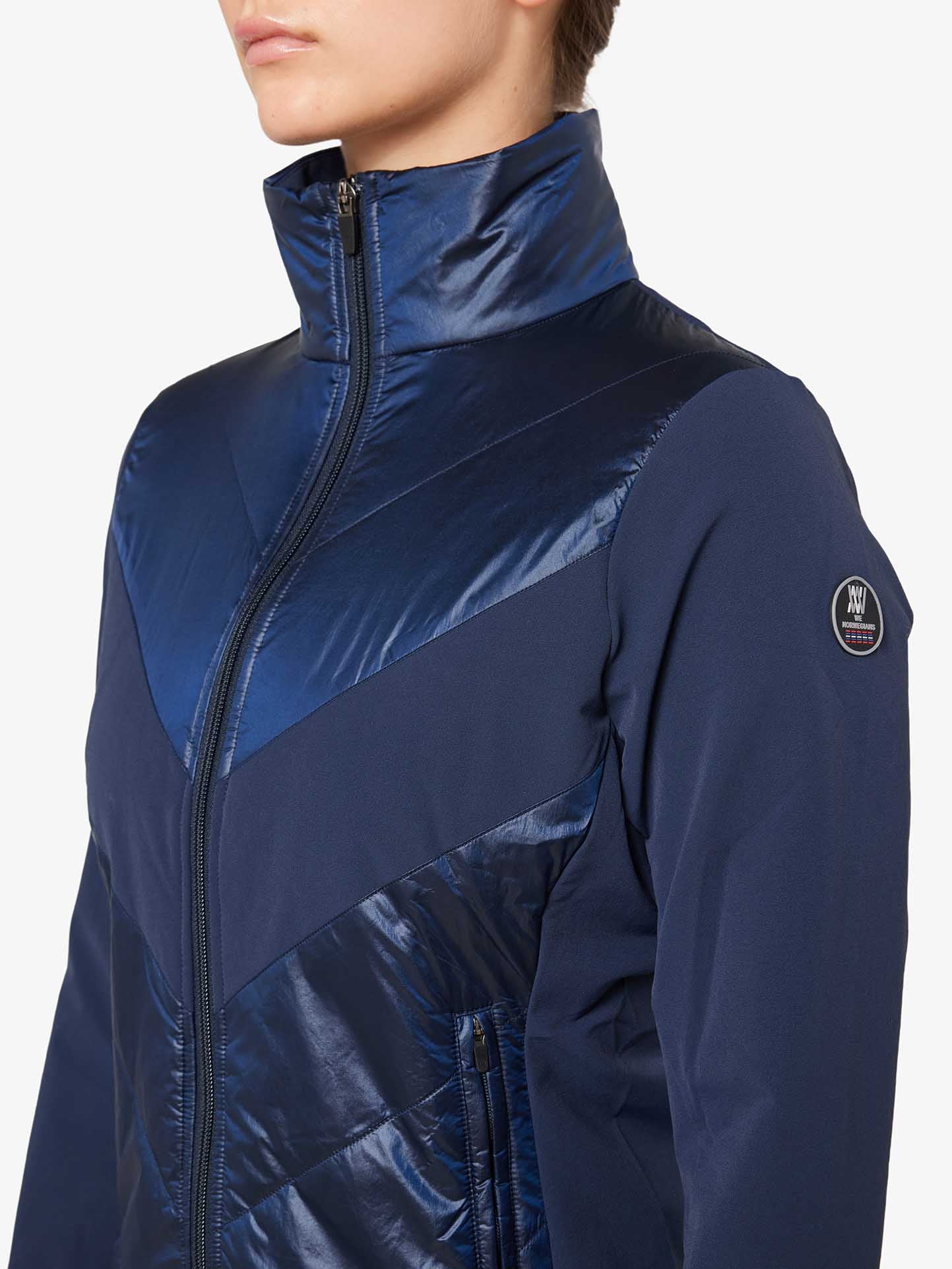 A person wearing a navy blue, long-sleeve We Norwegians Beito Jacket Women with a high collar. Designed for cross country skiing, the jacket showcases contrasting shiny and matte fabric panels. A circular patch with the logo is visible on the left sleeve. The background is plain white.