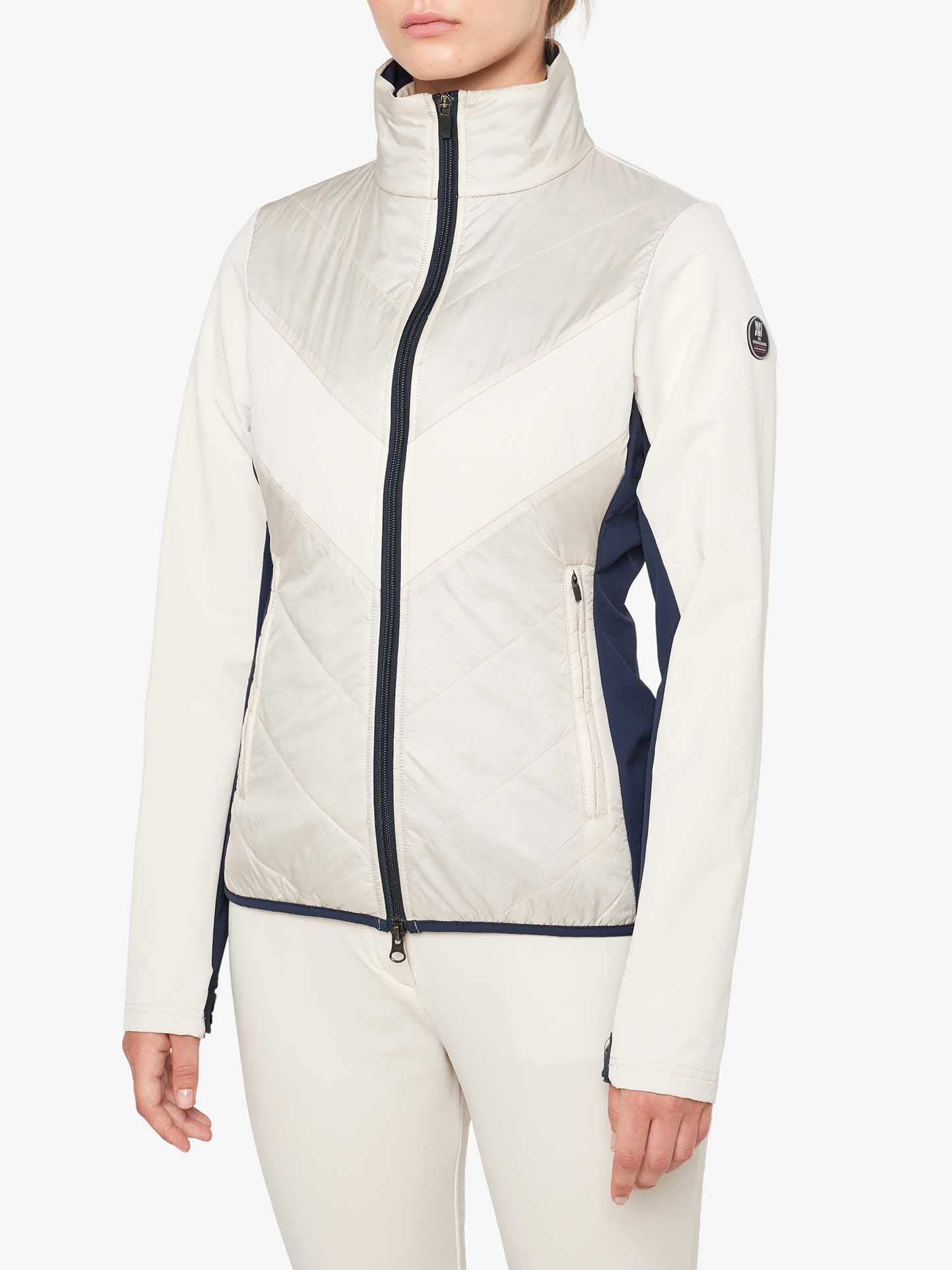 Person wearing a white and navy blue We Norwegians Beito Jacket Women with a high collar and chevron pattern on the front, standing against a plain background. The jacket, ideal for cross-country skiing, features a front zipper and two side pockets.