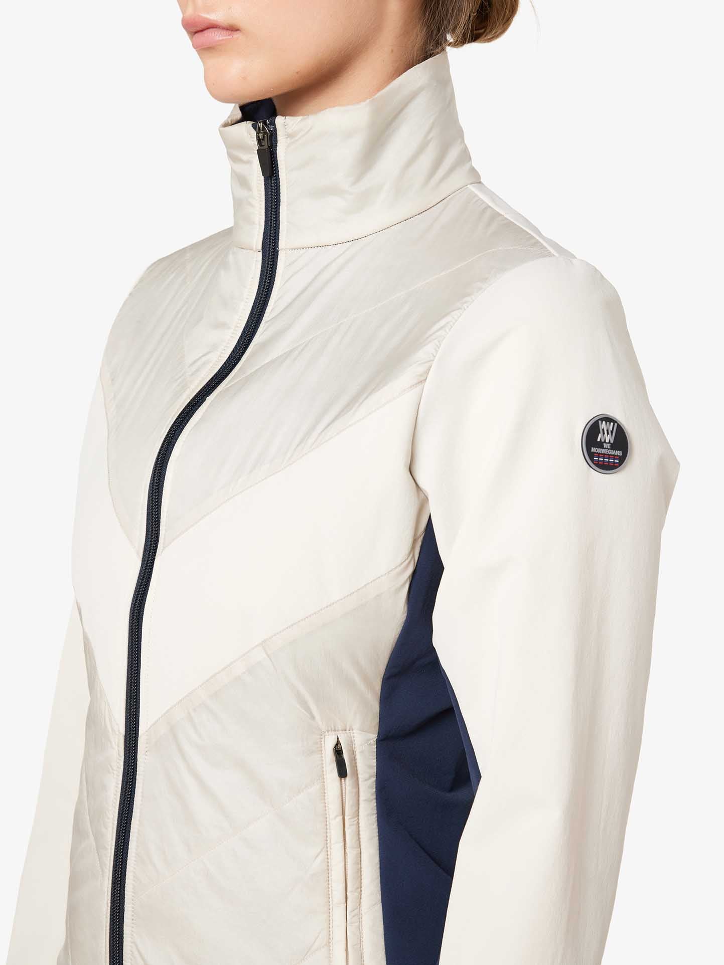 A person wearing the We Norwegians Beito Jacket Women, which is light-colored and high-collared with a front zipper. The jacket, designed for cross-country skiing, showcases a chevron pattern and a small circular logo patch on the upper sleeve. Only the person's upper torso and the side of their face are visible.