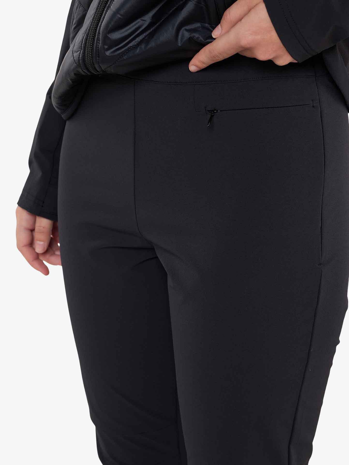 Close-up of a person wearing We Norwegians' Beito Pant Women, with a focus on the midsection and upper legs. A hand is partially visible, resting near a zippered pocket on the right side. The person is also wearing a dark top, and part of a crossbody bag appears at the top of the image.