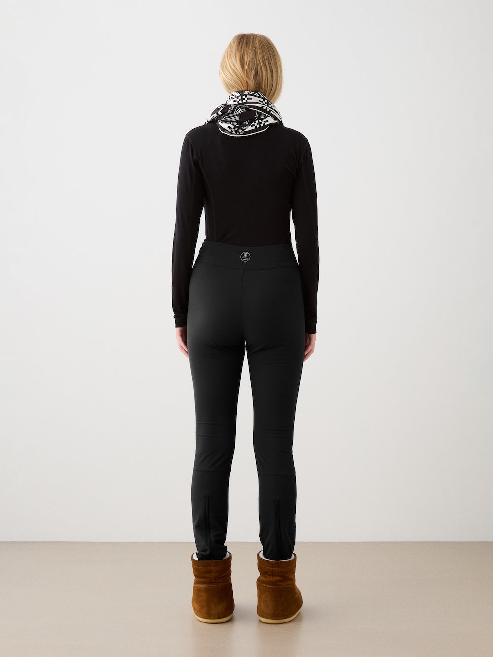 A woman with blonde hair, dressed in a black long-sleeve top, We Norwegians Beito Pant Women, brown boots, and a patterned scarf stands facing away from the camera against a plain background.