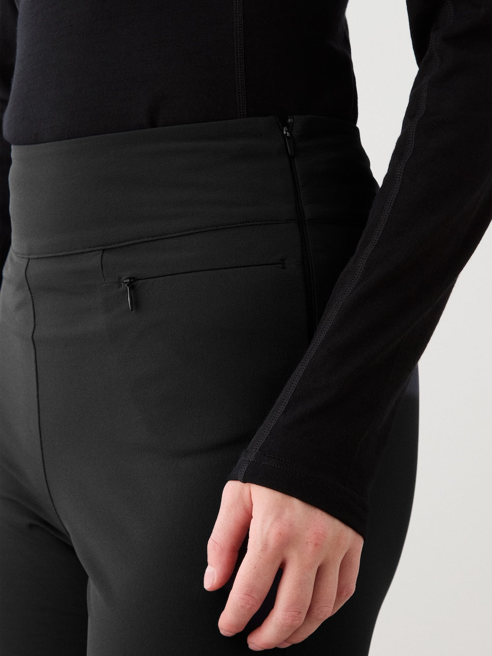 A person wears fitted black Beito Pant Women by We Norwegians with a long-sleeve top, hand near hip; a zipper and small pocket are visible on these stylish outdoor training pants.