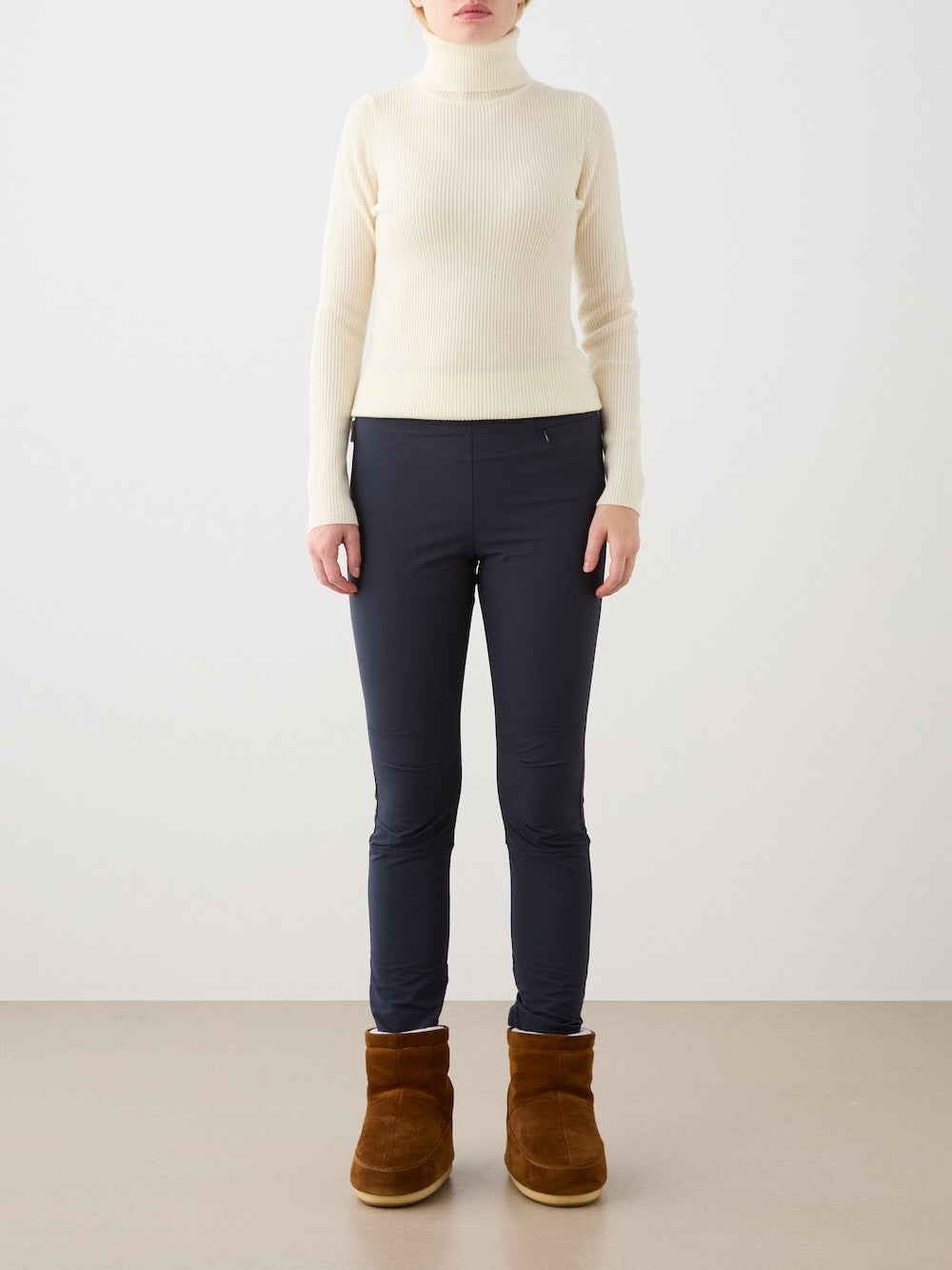 A person stands against a plain background wearing a cream turtleneck, We Norwegians Beito Pant Women, and brown ankle boots. Their arms are relaxed at their sides and their face is not visible.