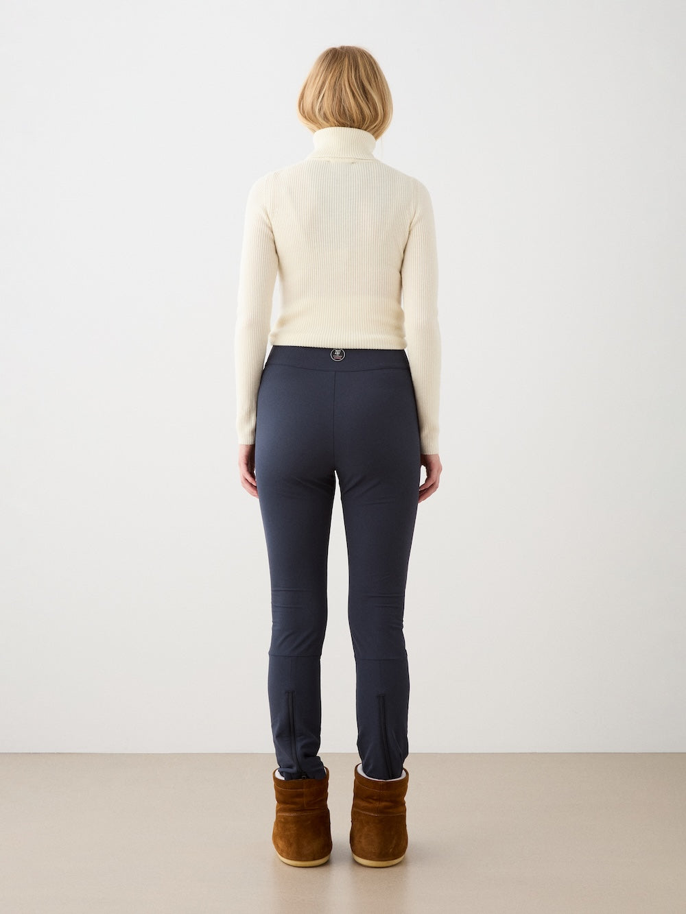 A woman with short blonde hair, dressed in a cream turtleneck sweater, We Norwegians Beito Pant Women in dark color, and brown boots, stands facing away against a plain light background.