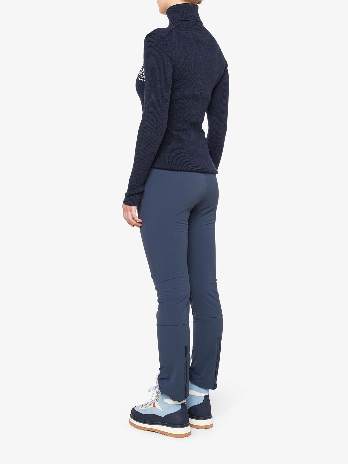 A person is seen standing sideways, wearing a dark navy long-sleeve sweater made from peached Merino wool, We Norwegians Beito Pant Women in blue, and light blue and navy hiking boots. The background is plain white. Only part of their face is visible, and their arms are by their sides.