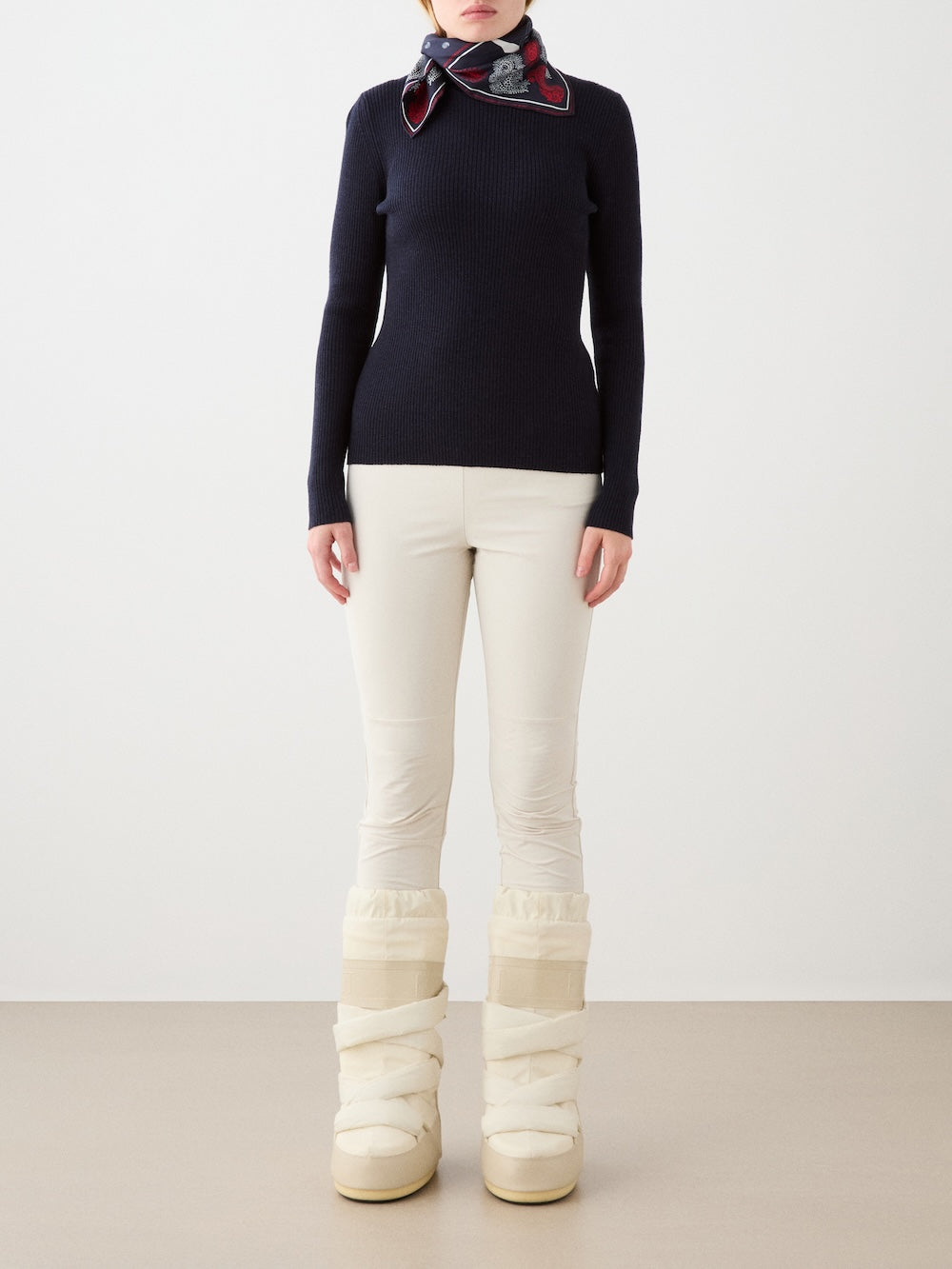A person stands against a plain background wearing a black long-sleeve sweater, a patterned scarf, cream We Norwegians Beito Pant Women outdoor training pants, and cream winter boots. Their face is partially out of frame.