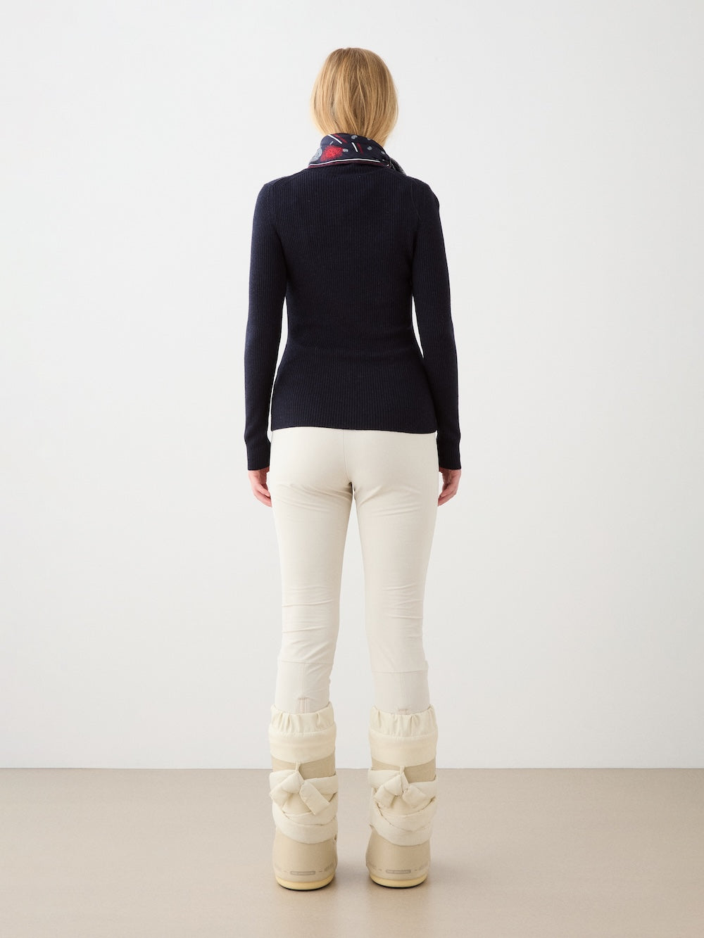 A person with blonde hair faces a white wall, wearing a dark long-sleeve top, We Norwegians Beito Pant Women in cream, a patterned scarf, and off-white winter boots.
