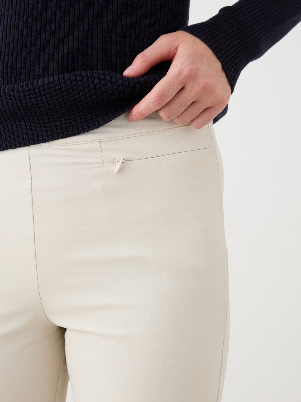 A person is shown from waist to mid-thigh wearing the We Norwegians Beito Pant Women in cream, featuring a small front pocket detail, and a navy ribbed sweater, with one hand resting on the waistband.