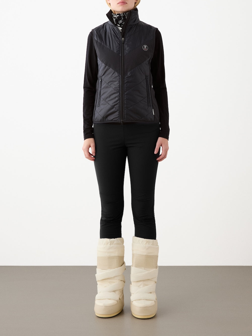 A person stands against a plain background wearing a black We Norwegians Beito Vest Women, black long-sleeve top, black pants, and cream padded winter boots. The vest offers non-fluorinated DWR for weather resistance. Their face is not visible.