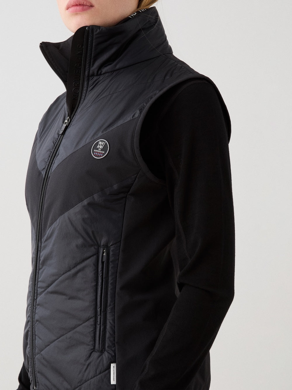 A woman wears the We Norwegians Beito Vest Women in black, featuring a high collar, non-fluorinated DWR finish, small chest logo, and zippered pocket over a black long-sleeve shirt. Her face is partially out of frame.