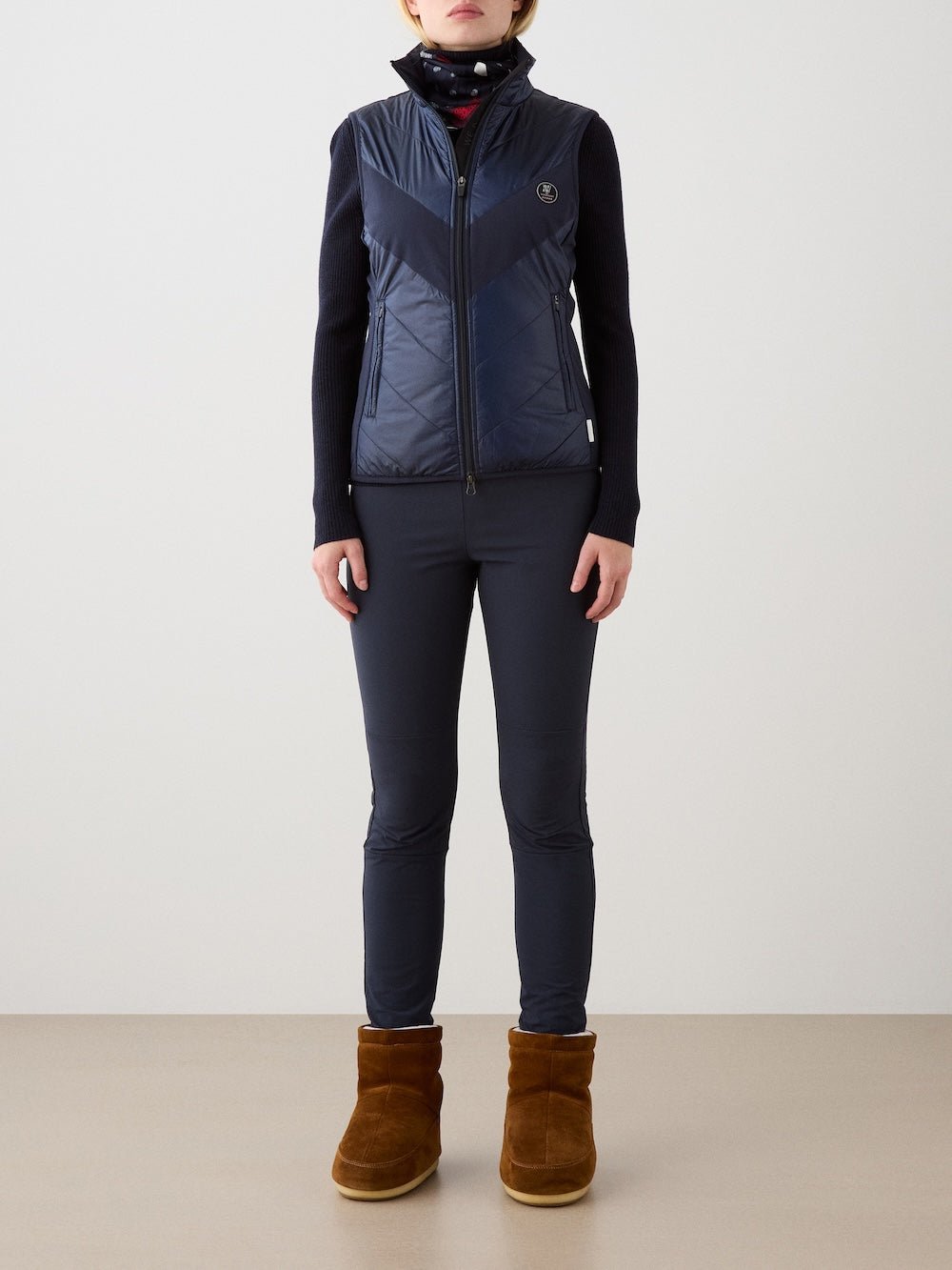 A person faces forward in a navy We Norwegians Beito Vest Women with non-fluorinated DWR, layered over a black long-sleeve top, navy pants, a neck scarf, and brown suede ankle boots, set against a plain light background.
