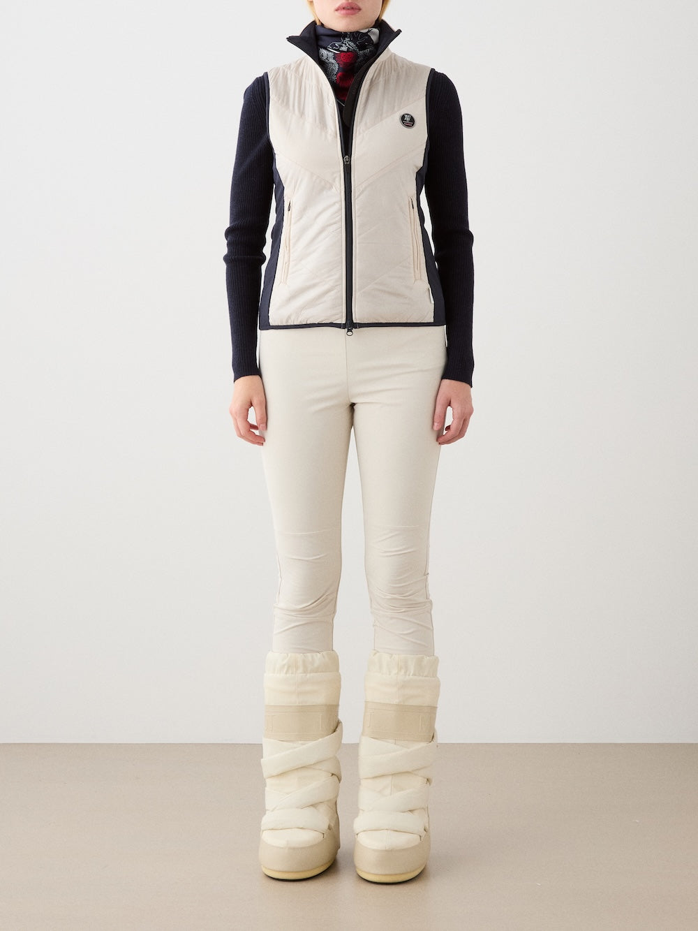 A person stands against a plain background wearing a light beige We Norwegians Beito Vest Women made from merino wool, paired with cream pants, beige puffy snow boots, a black long-sleeve top, and a patterned dark scarf. Their hands are at their sides.