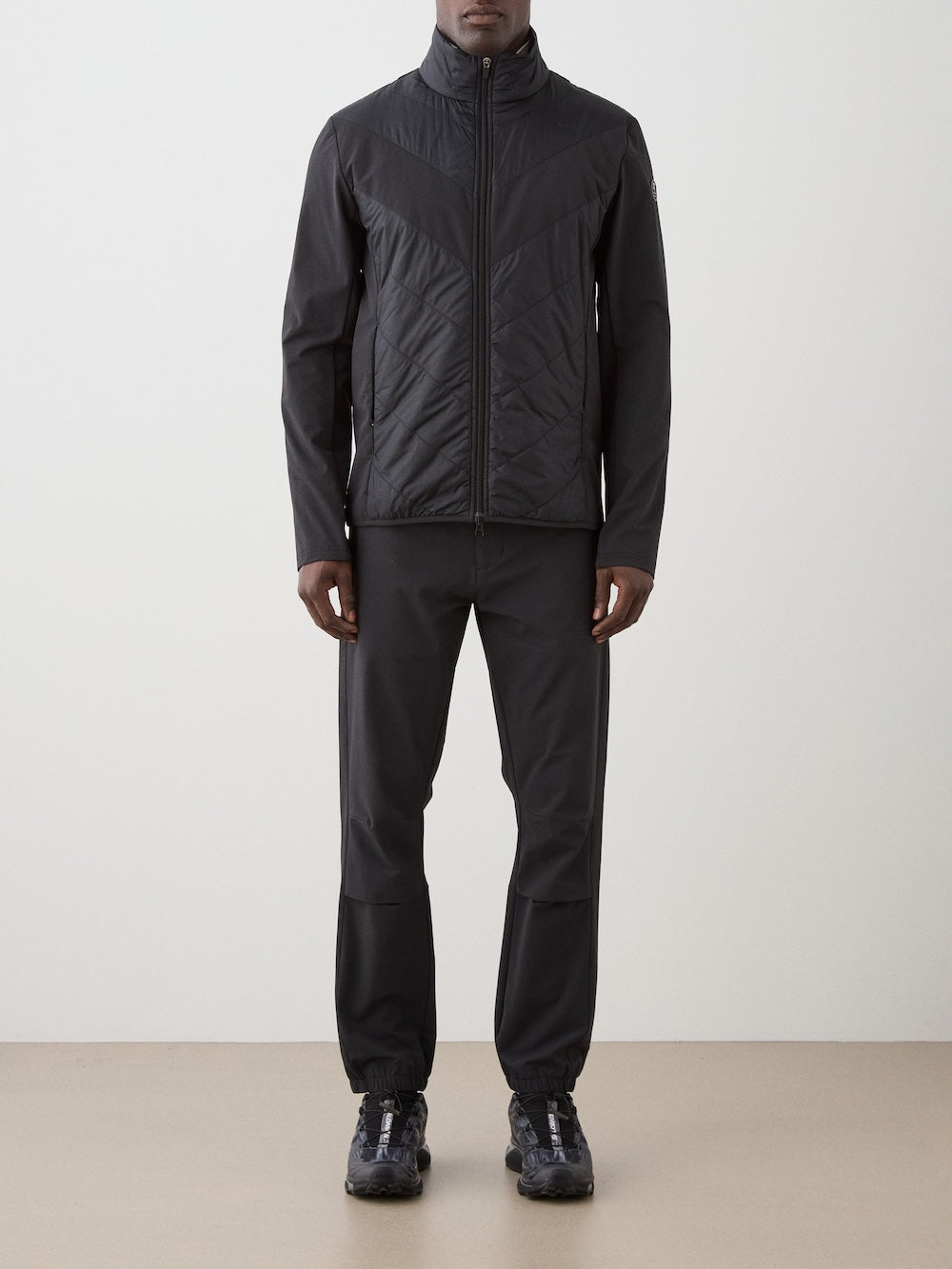 A person stands against a plain background wearing a black We Norwegians Beito Jacket Men—an eco-friendly, water-resistant zip-up—paired with black pants and black athletic shoes. Their face is not visible, and their arms are relaxed at their sides.