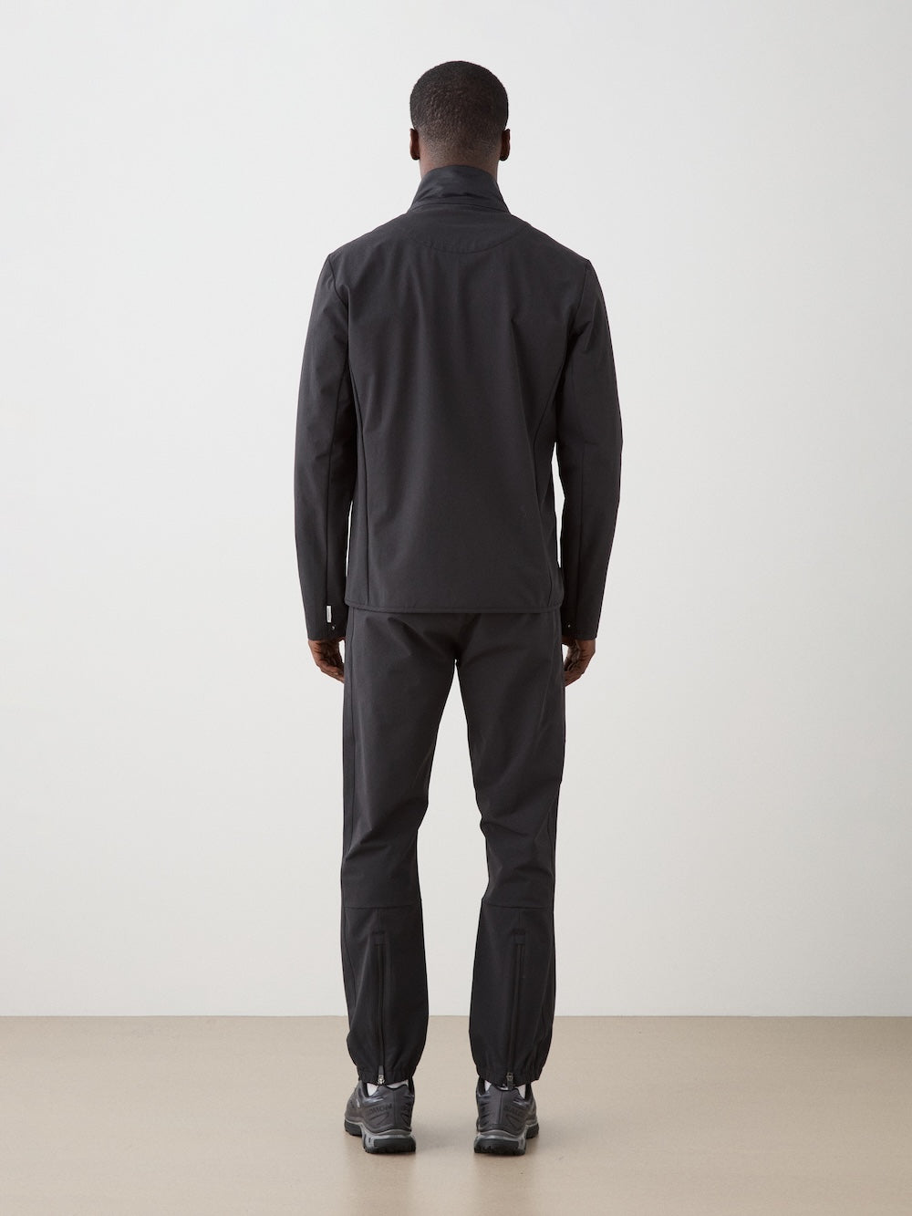 A man faces away from the camera, wearing the We Norwegians Beito Jacket Men—an eco-friendly, water-resistant merino wool jacket—paired with black pants and sneakers against a plain light backdrop.