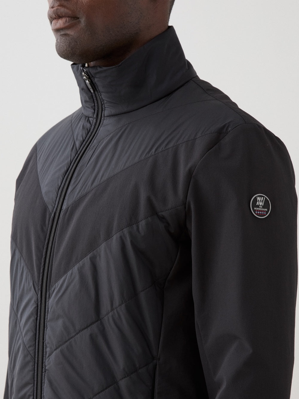 A man wears the We Norwegians Beito Jacket Men in black, featuring a high collar, quilted details, eco-friendly water-resistant finish, and a small circular logo patch on the upper sleeve. The background is plain and light-colored.