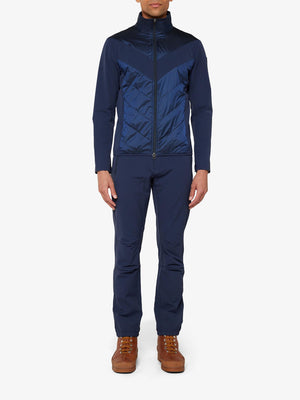 A person stands facing forward, wearing a dark blue Beito Jacket Men by We Norwegians featuring a chevron pattern, along with matching pants. The merino wool outfit is complemented by brown hiking boots, all set against a white background.