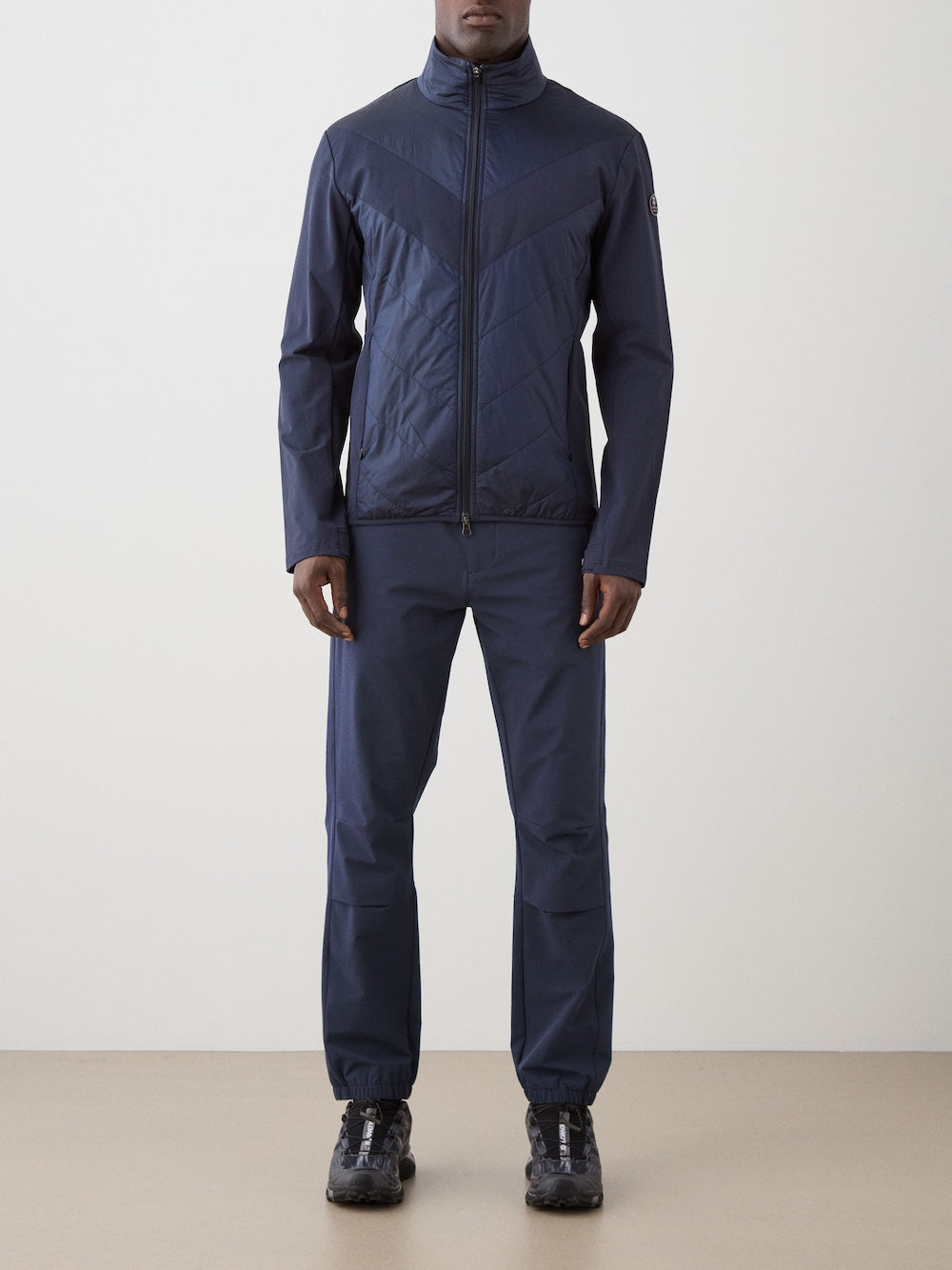 A person stands against a plain background wearing the We Norwegians Beito Jacket Men in navy merino wool with matching navy pants and black sneakers—ideal for cold-weather adventures.