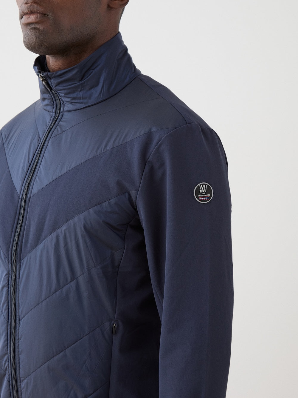 A man wears a navy blue We Norwegians Beito Jacket Men—a water-resistant, eco-friendly zip-up with a high collar and logo patch on the upper sleeve—standing against a plain light background.