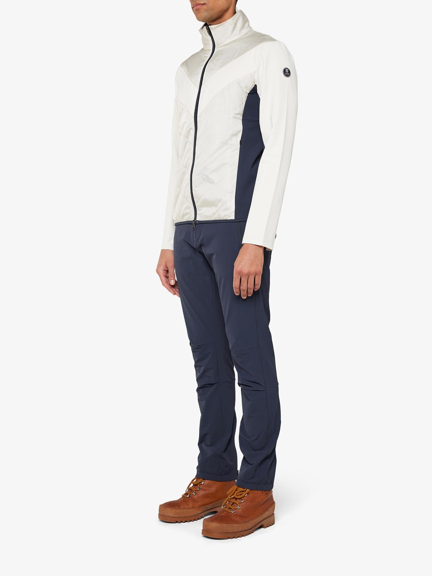 A man is standing in a Beito Jacket Men by We Norwegians, which is white and navy, paired with navy pants. He is wearing brown hiking boots and stands against a plain white background. The jacket, perfect for cross-country skiing, complements his outfit effortlessly while ensuring comfort with its merino wool fabric. His face is not visible in the image.