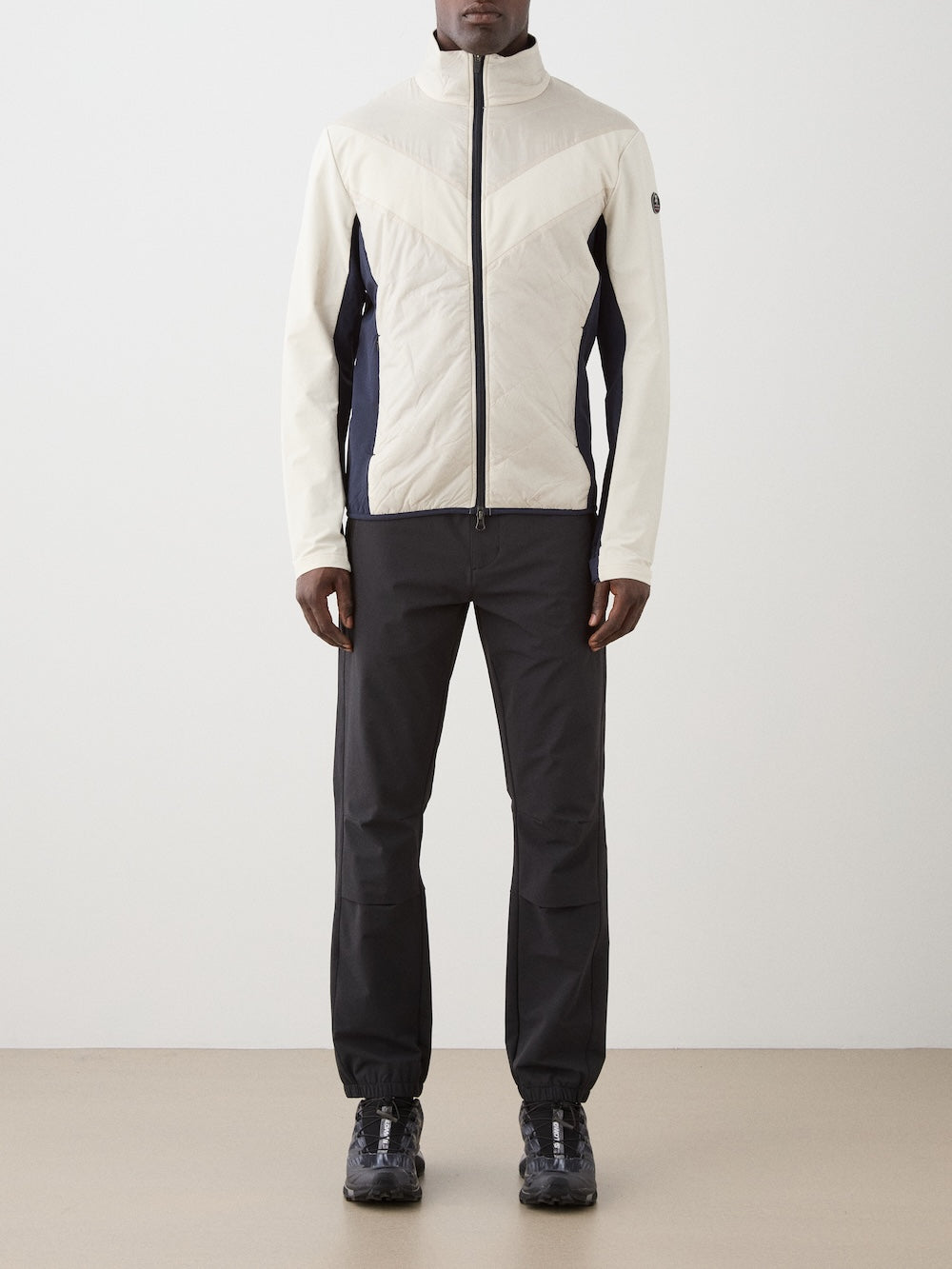 A person stands against a plain background wearing the We Norwegians Beito Jacket Men in cream and black, paired with black pants and sneakers. Their hands hang by their sides, face unseen. This eco-friendly, water-resistant jacket adds modern style.