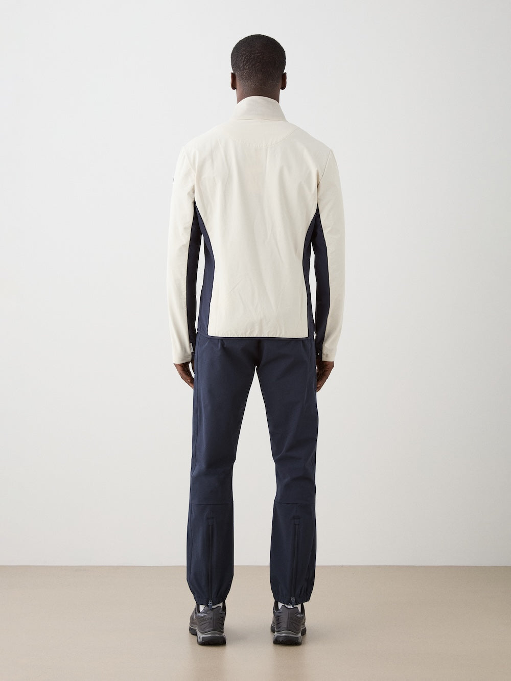 A person stands facing away, wearing the We Norwegians Beito Jacket Men—an eco-friendly, water-resistant jacket with dark sleeves—paired with dark pants in a minimal indoor setting with a beige floor and light gray wall.