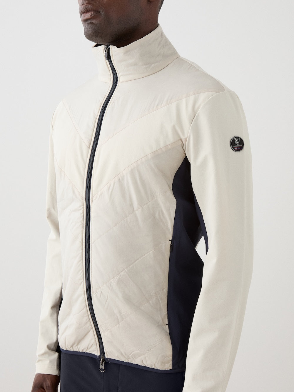 A man stands against a light background wearing the We Norwegians Beito Jacket Men in light beige with dark side panels, a chevron quilted pattern, and round black sleeve logo. This eco-friendly, water-resistant jacket blends style and functionality.