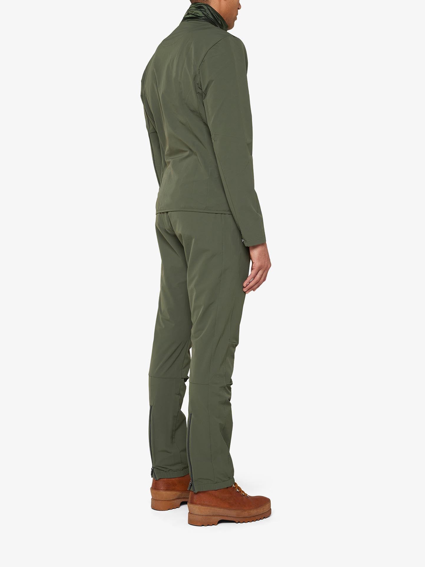 A person is standing with their back to the camera, dressed in an olive green Beito Jacket Men by We Norwegians and matching pants. They are also wearing brown ankle boots against a plain white background.