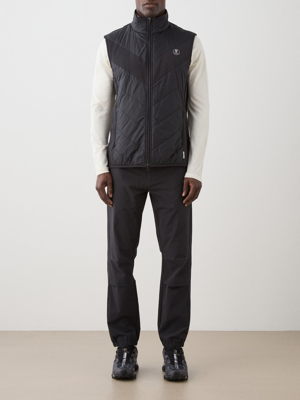A person stands indoors wearing the We Norwegians Beito Vest Men in black over a white long-sleeve shirt, with black pants and sneakers, against a plain background—an effortless approach to performance layering.