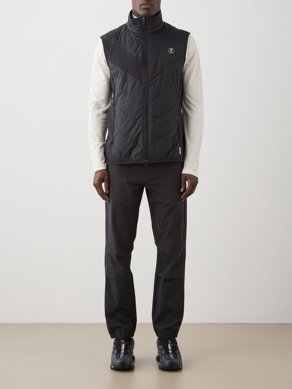 A person stands against a plain background wearing a white long-sleeve shirt, black pants with elastic cuffs, black sneakers, and the We Norwegians Beito Vest Men—a black quilted layer with a small round logo on the left chest.