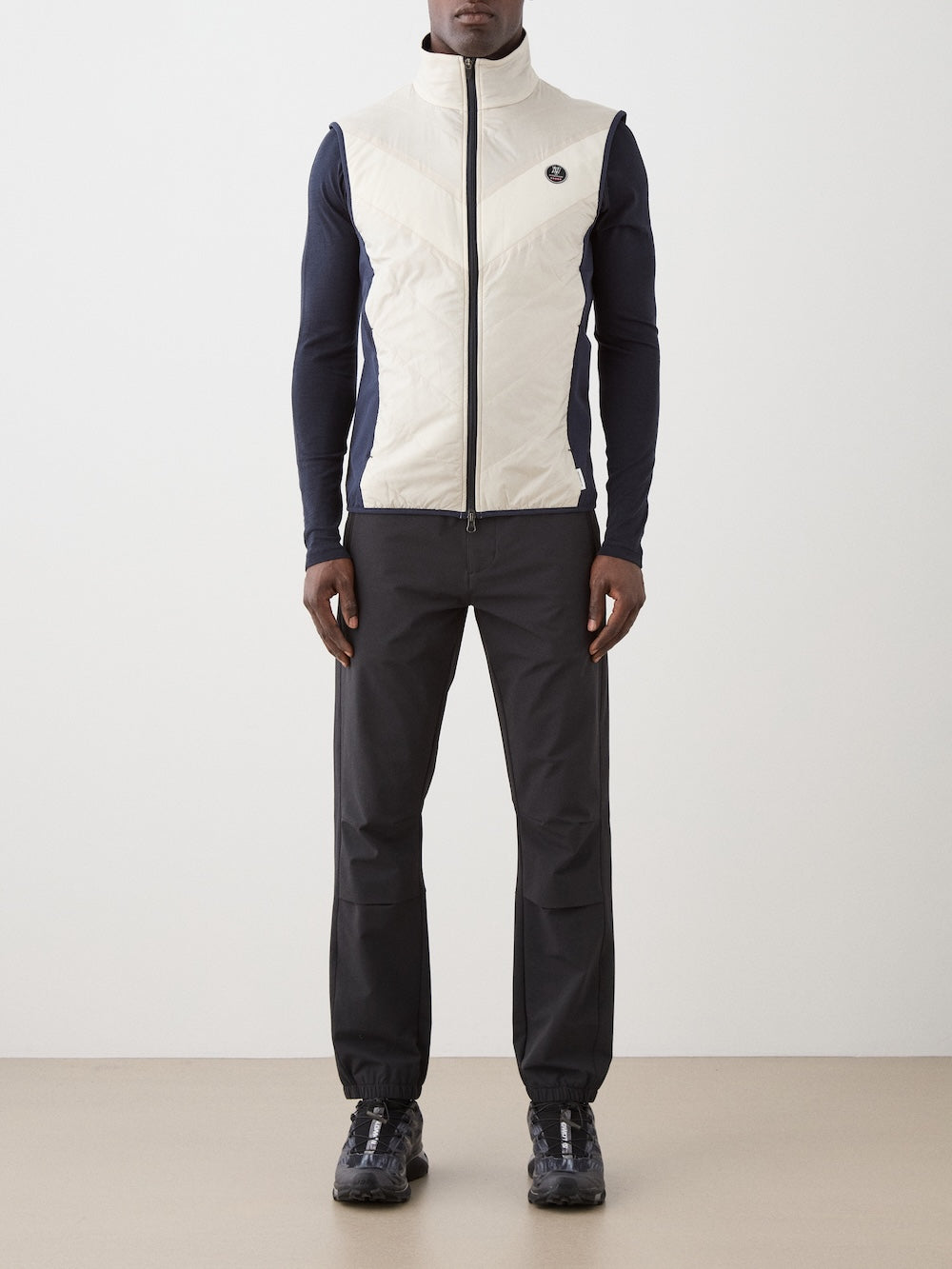 A man stands facing forward, wearing a white We Norwegians Beito Vest Men layered over a navy long-sleeve shirt, black pants with elastic cuffs, and black sneakers, set against a plain light background.