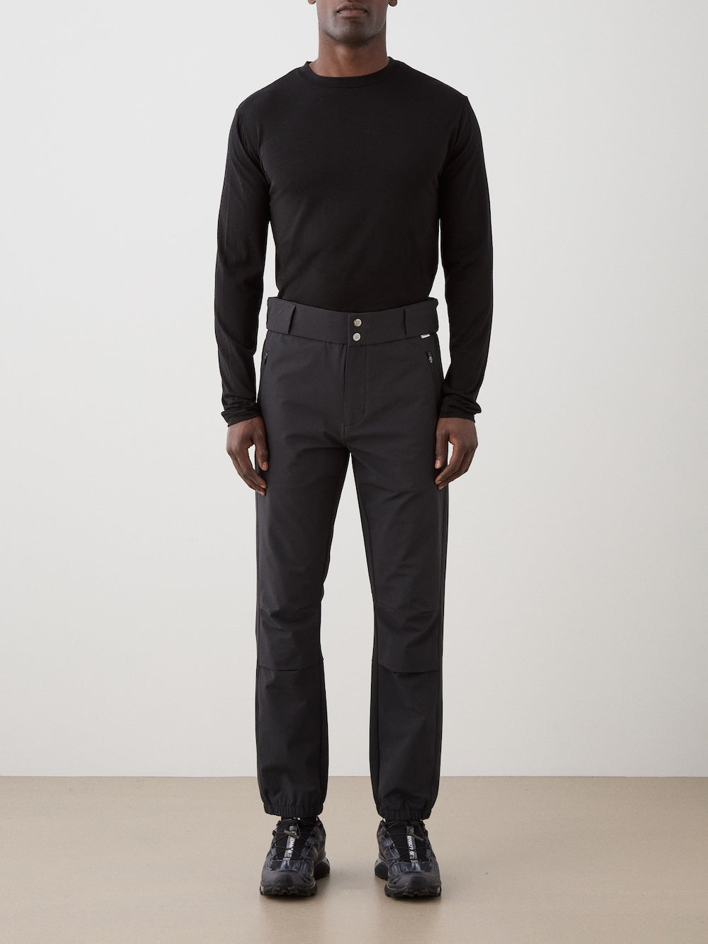 A person stands against a plain background wearing a black long-sleeve shirt, We Norwegians Beito Pant Men with belt loops and elasticized cuffs, and black athletic shoes for a simple, monochromatic look.
