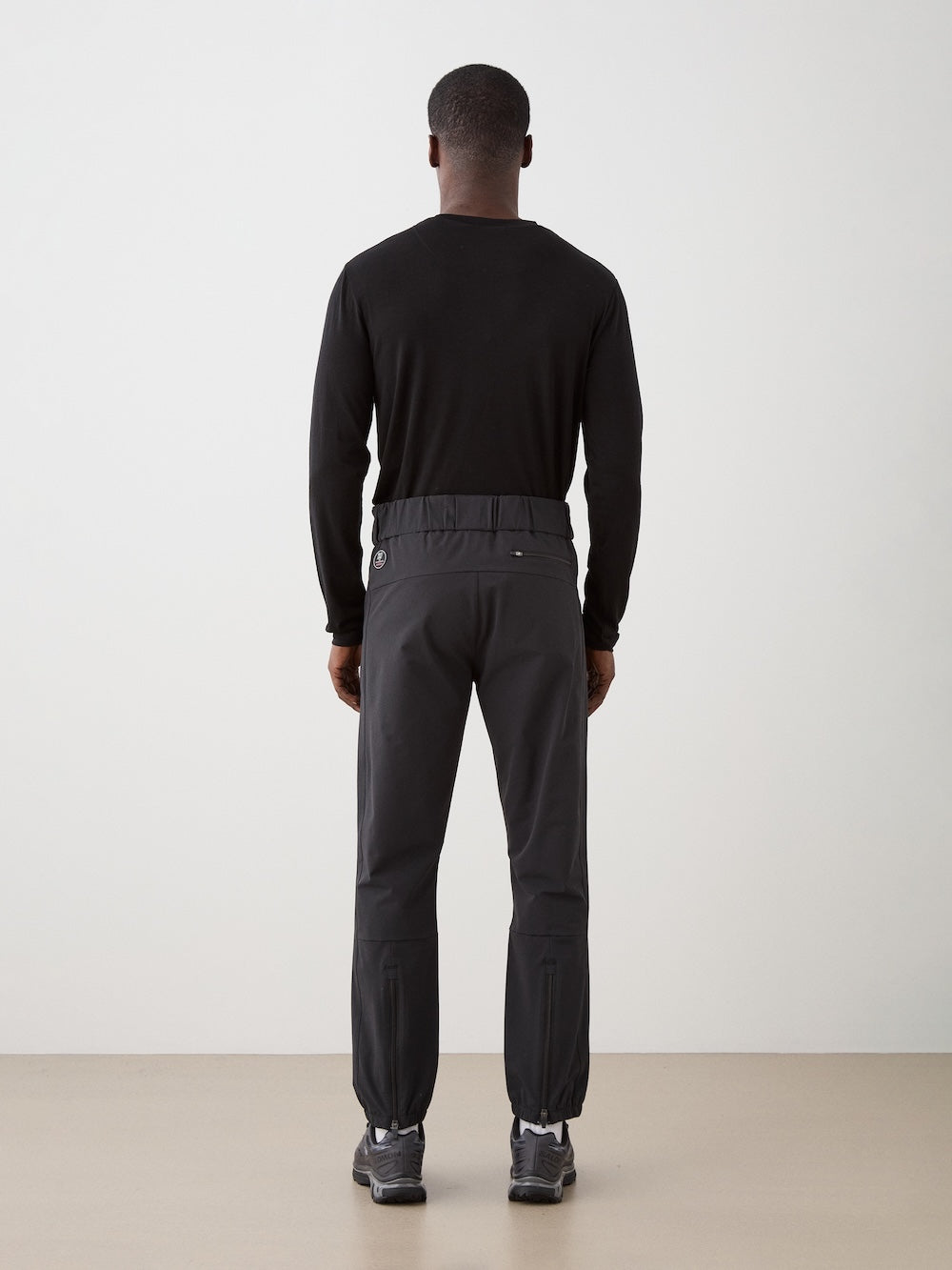 A man stands with his back to the camera, wearing a black long-sleeve shirt, We Norwegians Beito Pant Men with elastic cuffs, and gray sneakers against a plain light background.