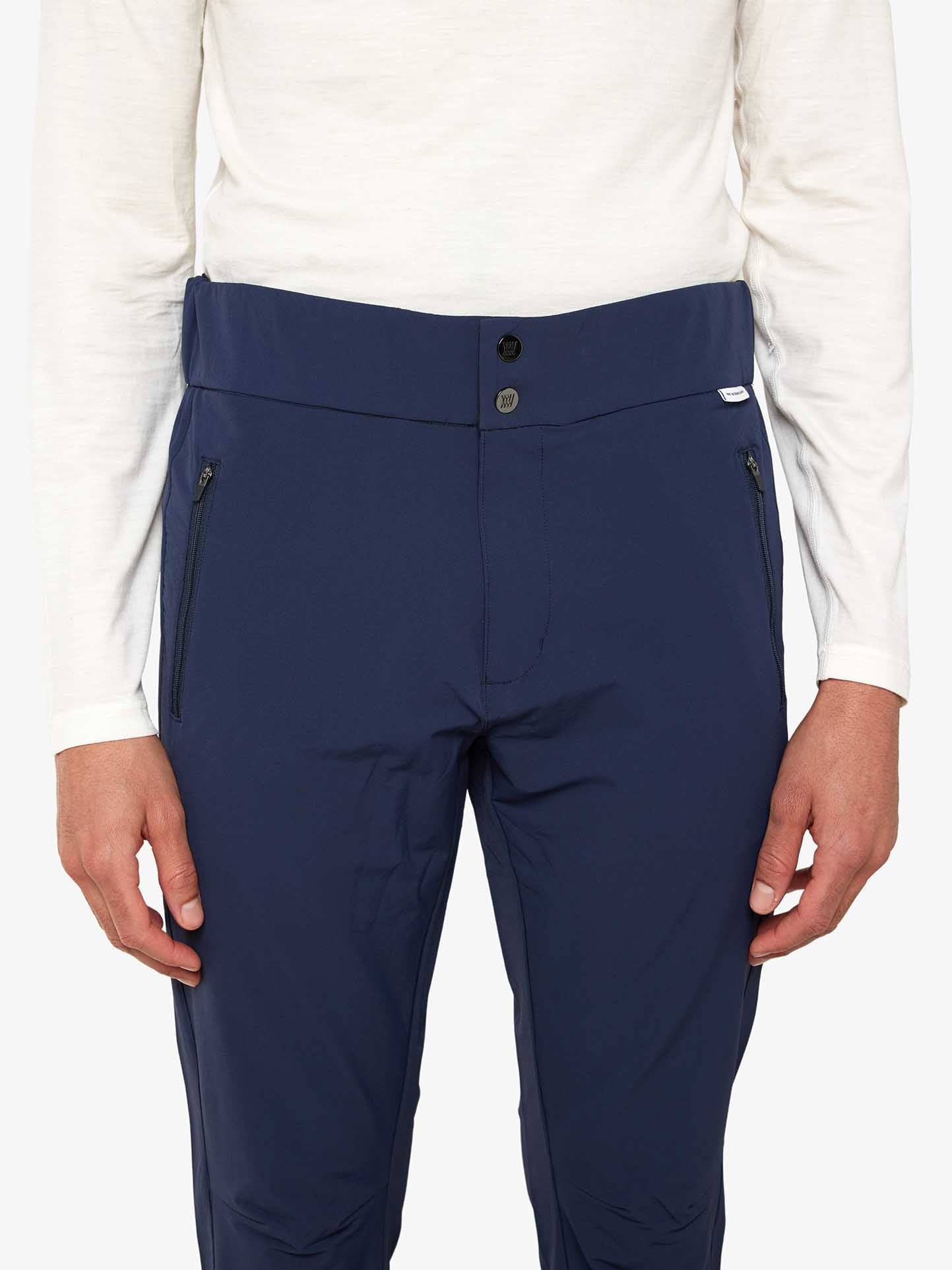 A person wearing a plain white long-sleeved shirt and the Beito Pant Men from We Norwegians, in dark blue with a button and zipper closure. These pants are made of breathable stretchy quality fabric, featuring zippered pockets on each side and an adjustable leg opening. The image is cropped to show only the person's torso and legs against a plain background.