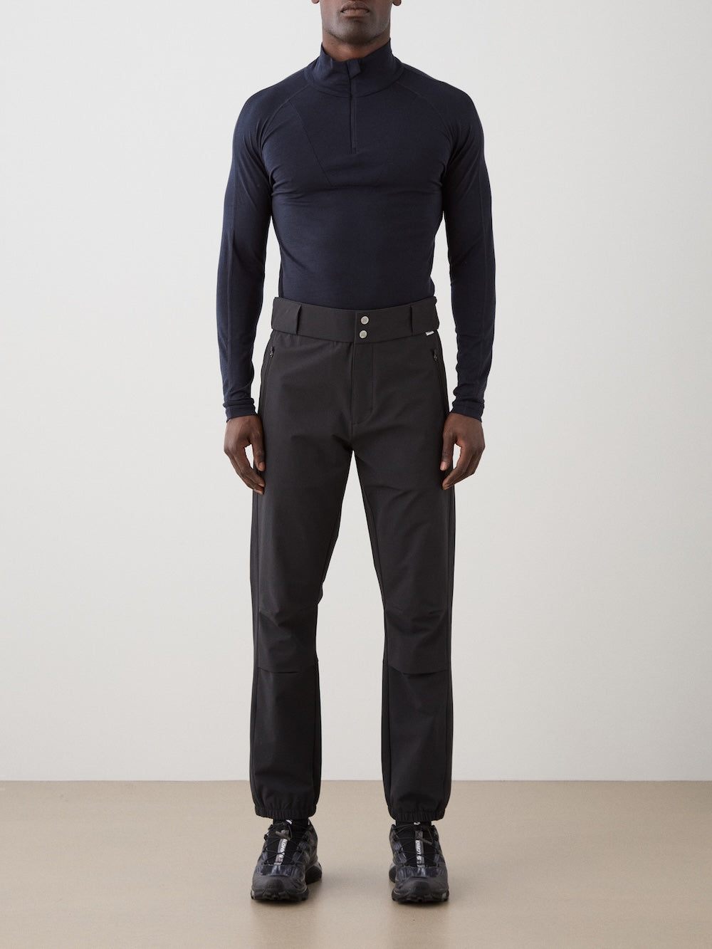 A person stands facing forward against a plain background, wearing a fitted long-sleeve black top, We Norwegians Beito Pant Men in black with a belt, and black athletic shoes.