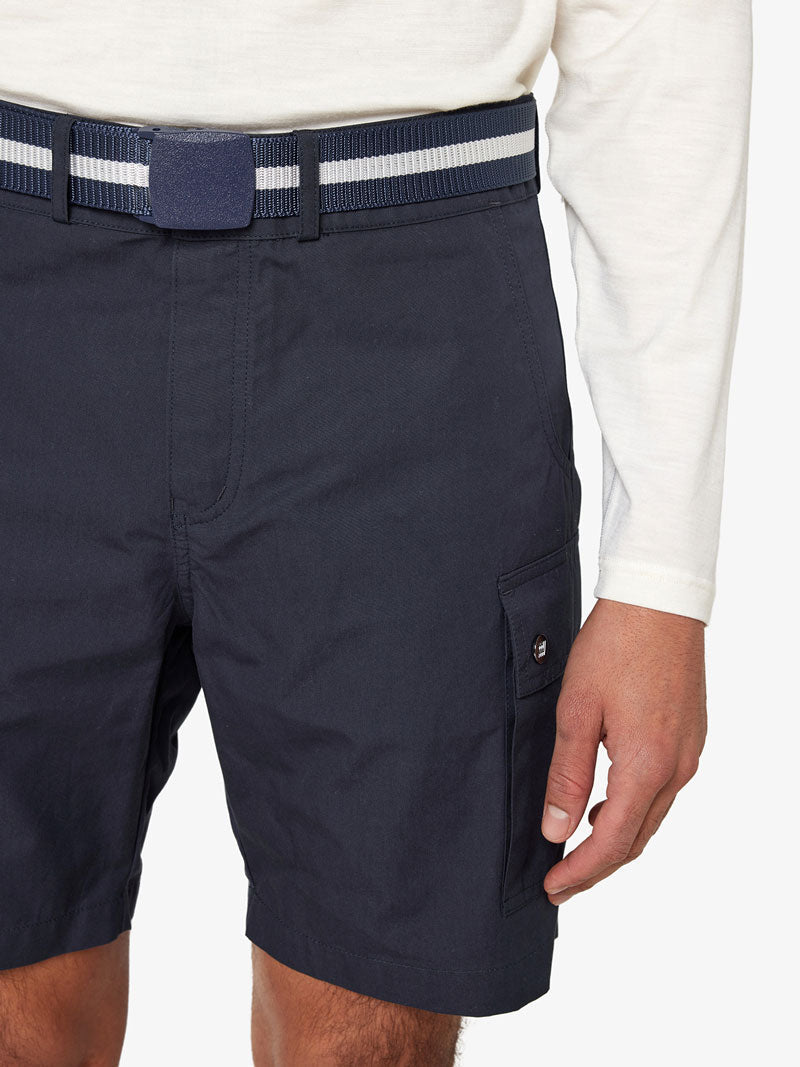 A person wearing a white long-sleeve shirt and navy blue belted cargo shorts with the Vidda Belt by We Norwegians, featuring an adjustable belt and side pocket. Only the lower part of the shirt, upper part of the shorts, and hand are visible against a plain background, highlighting the unisex accessory.