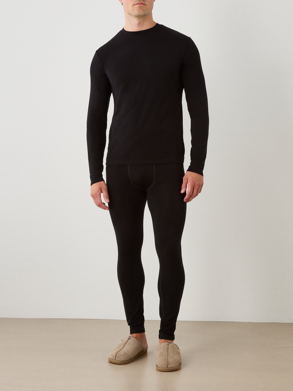 A man wearing the We Norwegians Sno Merino Long Sleeve Men and matching leggings stands against a plain background with his hands at his sides, also wearing light brown slippers.