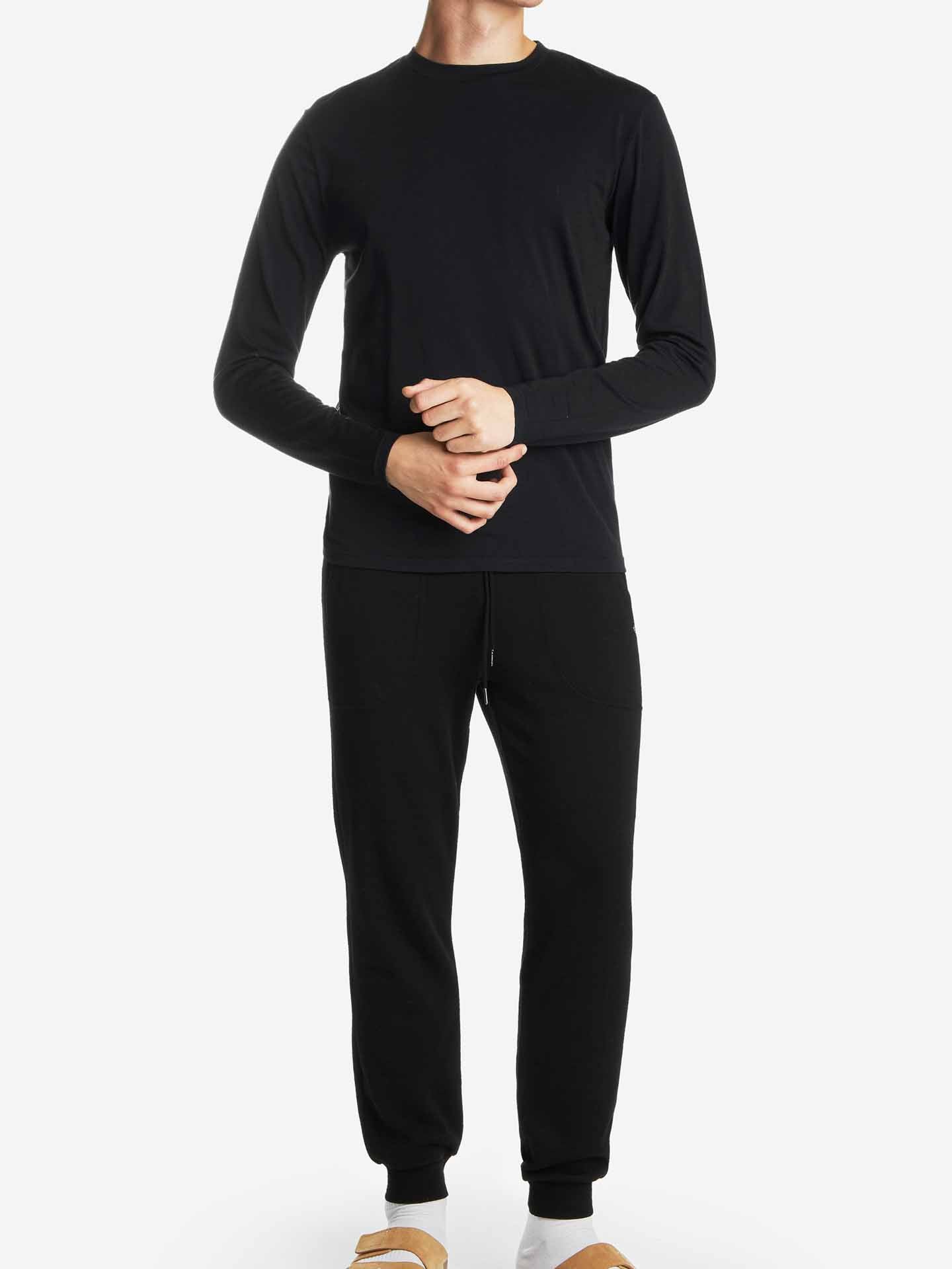 A person is wearing the Snø Merino Long Sleeve Men shirt by We Norwegians, along with matching pants, against a white background. Their hands are clasped in front, highlighting the comfort of the stretchy fabric. Brown slippers are visible on their feet, though their head is out of view.