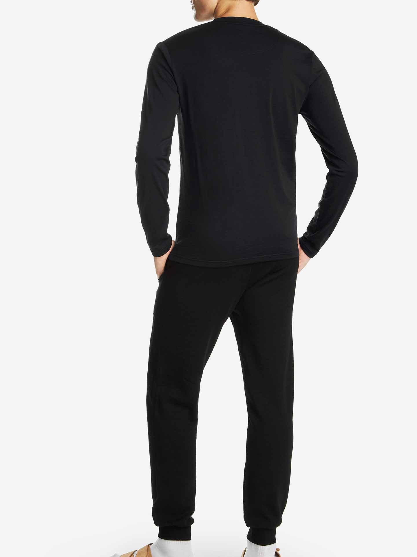 A person is standing with their back to the camera, dressed in the Snø Merino Long Sleeve Men shirt from We Norwegians, paired with matching stretchy pants. They are also wearing socks and sandals against a plain white background.