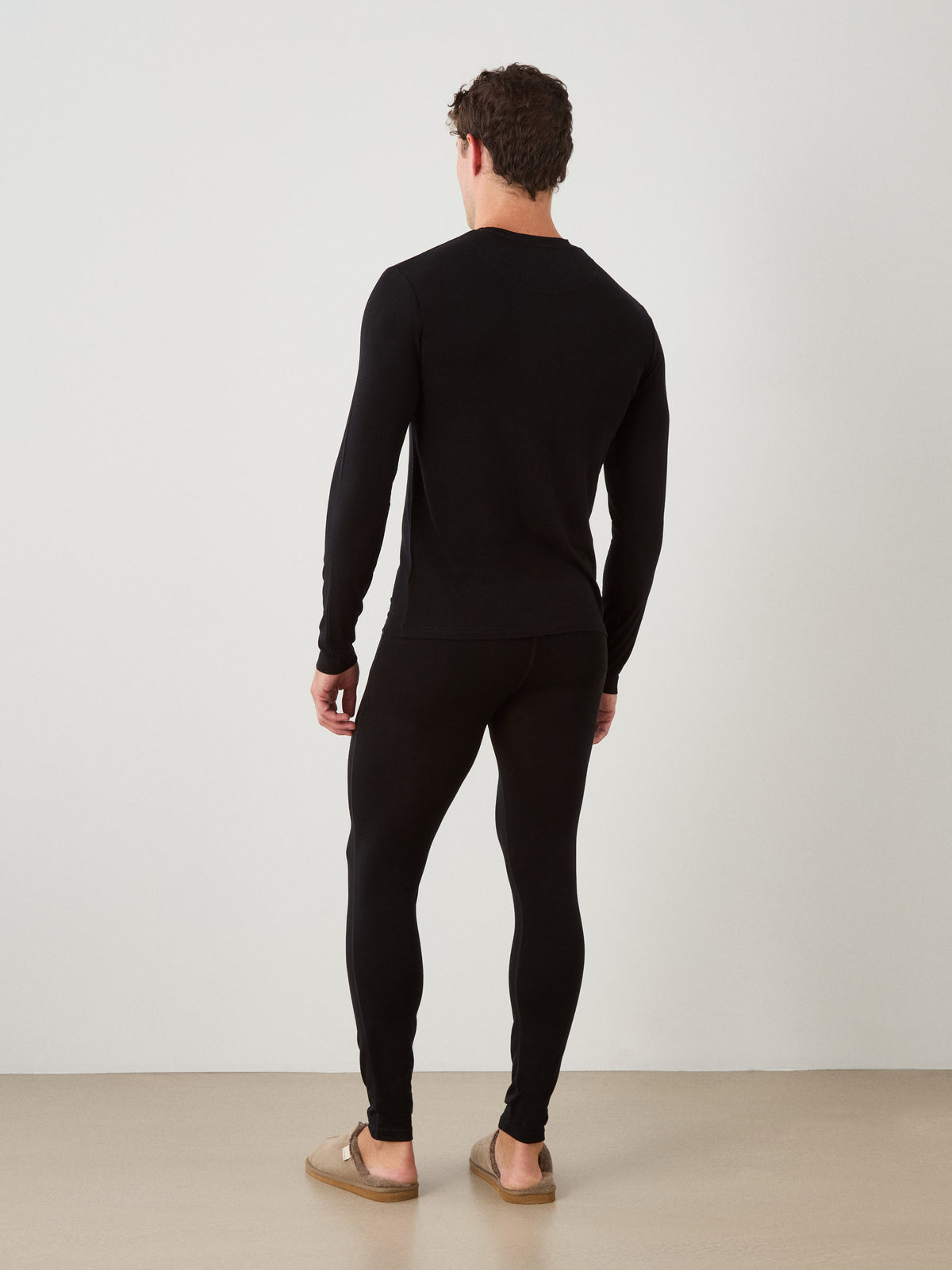 A man faces away, wearing the We Norwegians Sno Merino Long Sleeve Men in black and matching leggings with tan slip-on shoes, set against a plain white background.