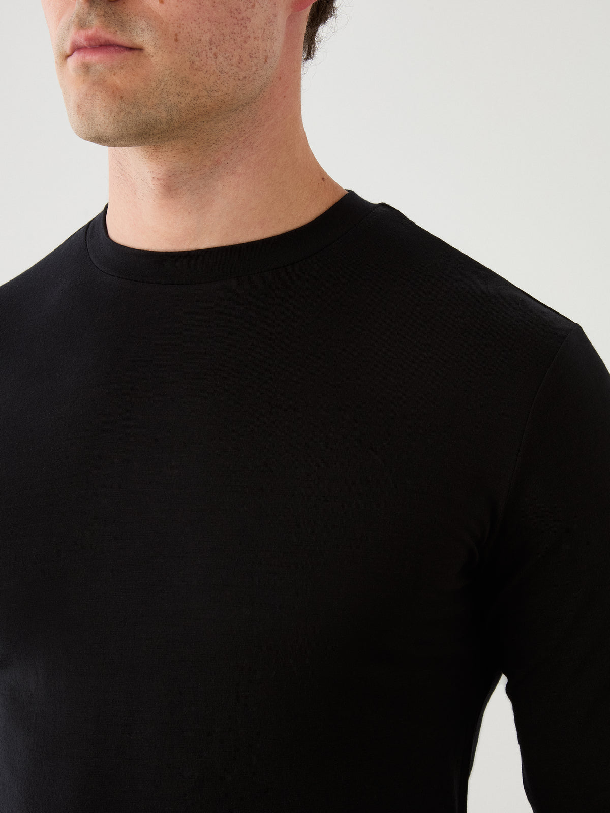 A man wearing the We Norwegians Sno Merino Long Sleeve Men in plain black is shown from the shoulders up against a white background, his face partially visible and turned slightly to the left.