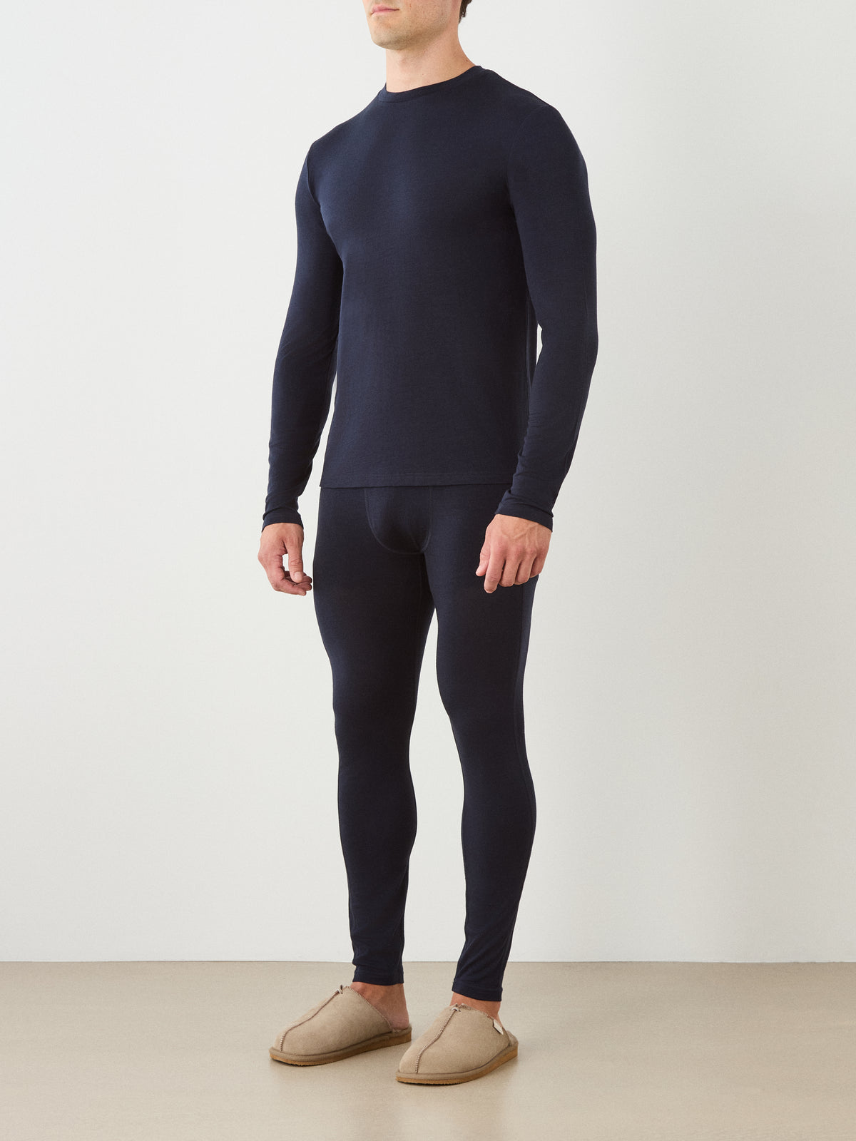 A man stands by a plain wall wearing the We Norwegians Sno Merino Long Sleeve Men in navy blue with matching leggings and beige slippers.