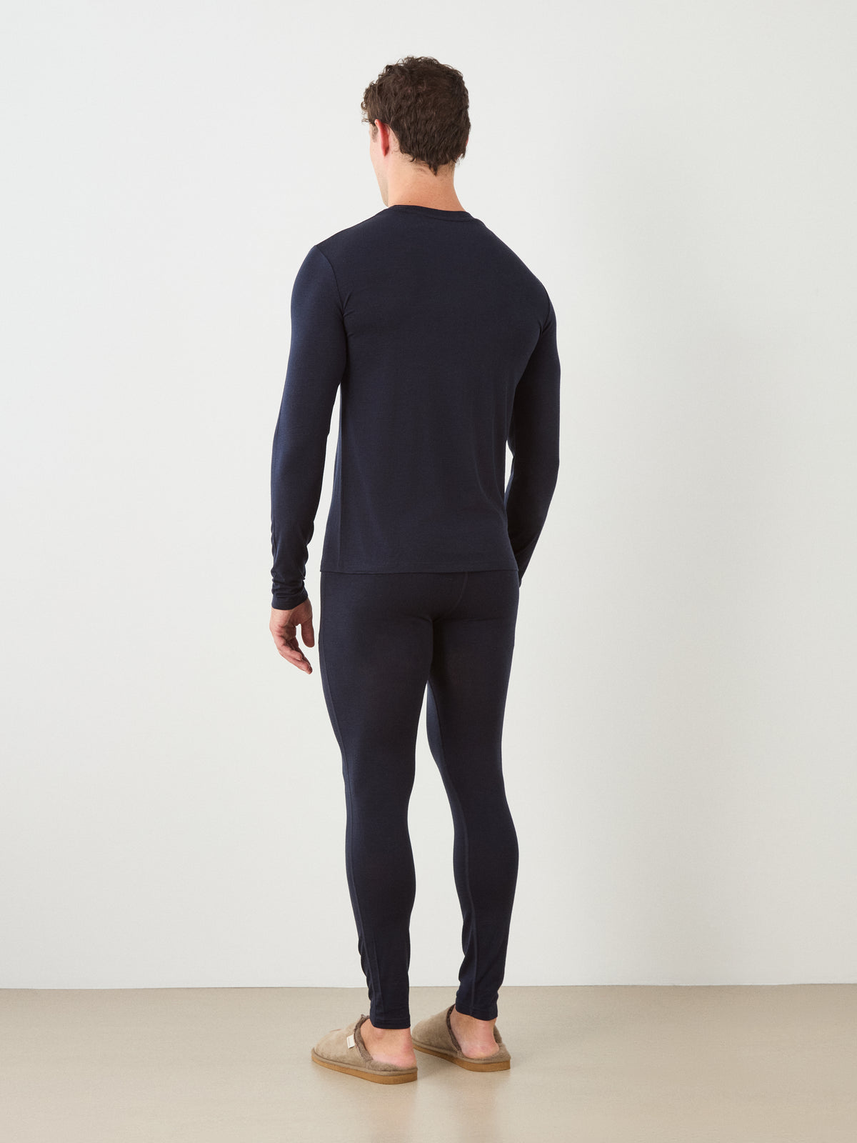 A man faces away, wearing the We Norwegians Sno Merino Long Sleeve Men and matching leggings with brown slippers, set against a plain white wall and beige floor.
